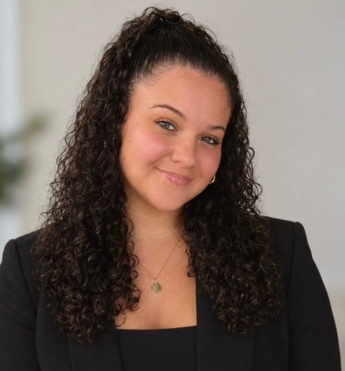 A young woman with curly dark hair styled in a high ponytail, wearing a black blazer and a gold necklace, smiling in an indoor setting.