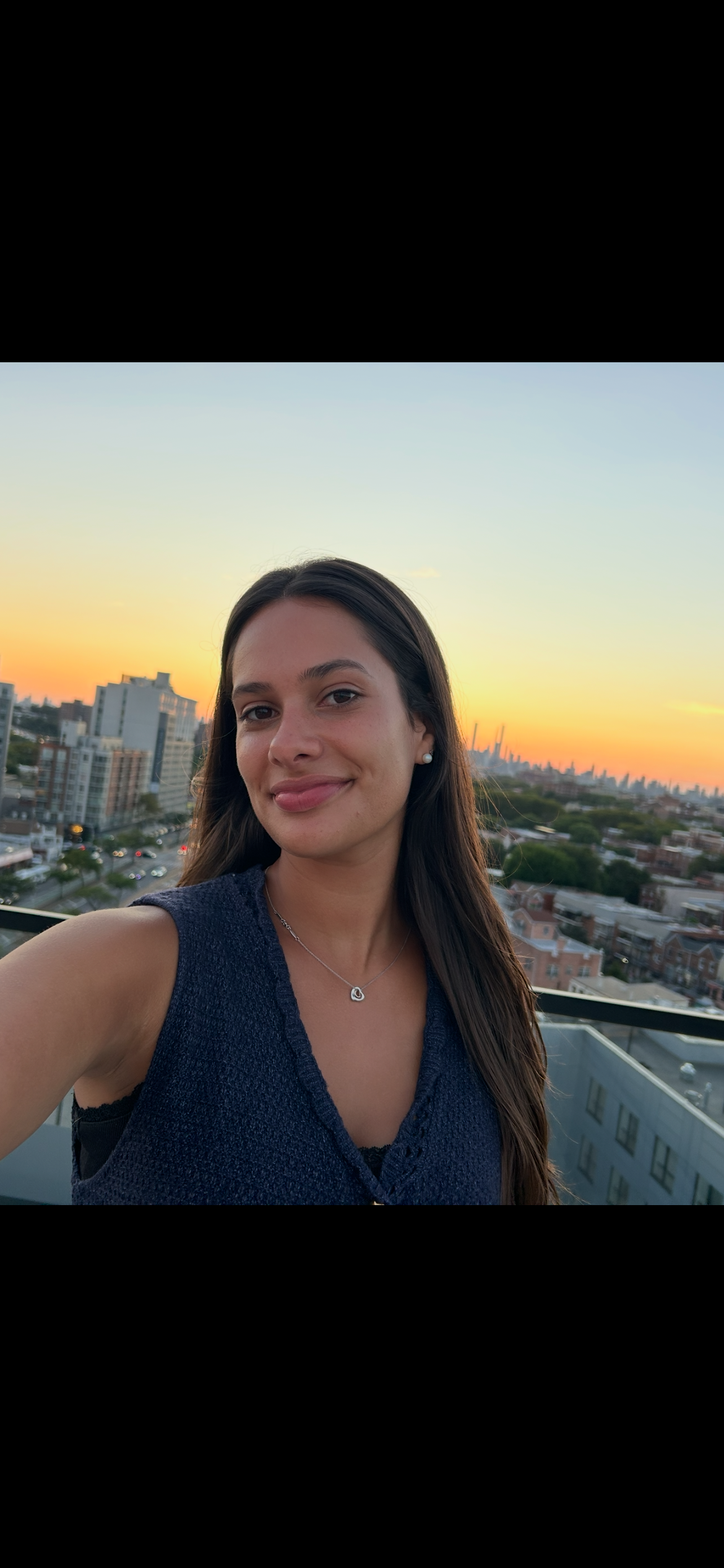 Young woman taking a selfie on a rooftop with a city skyline at sunset in the background.