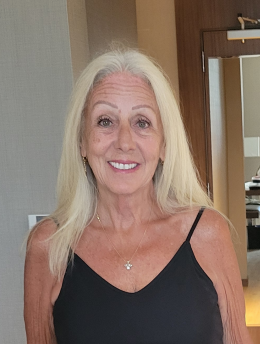 A smiling elderly woman with long, gray hair, wearing a black spaghetti strap top and a necklace, standing indoors near a mirror and doorframe.