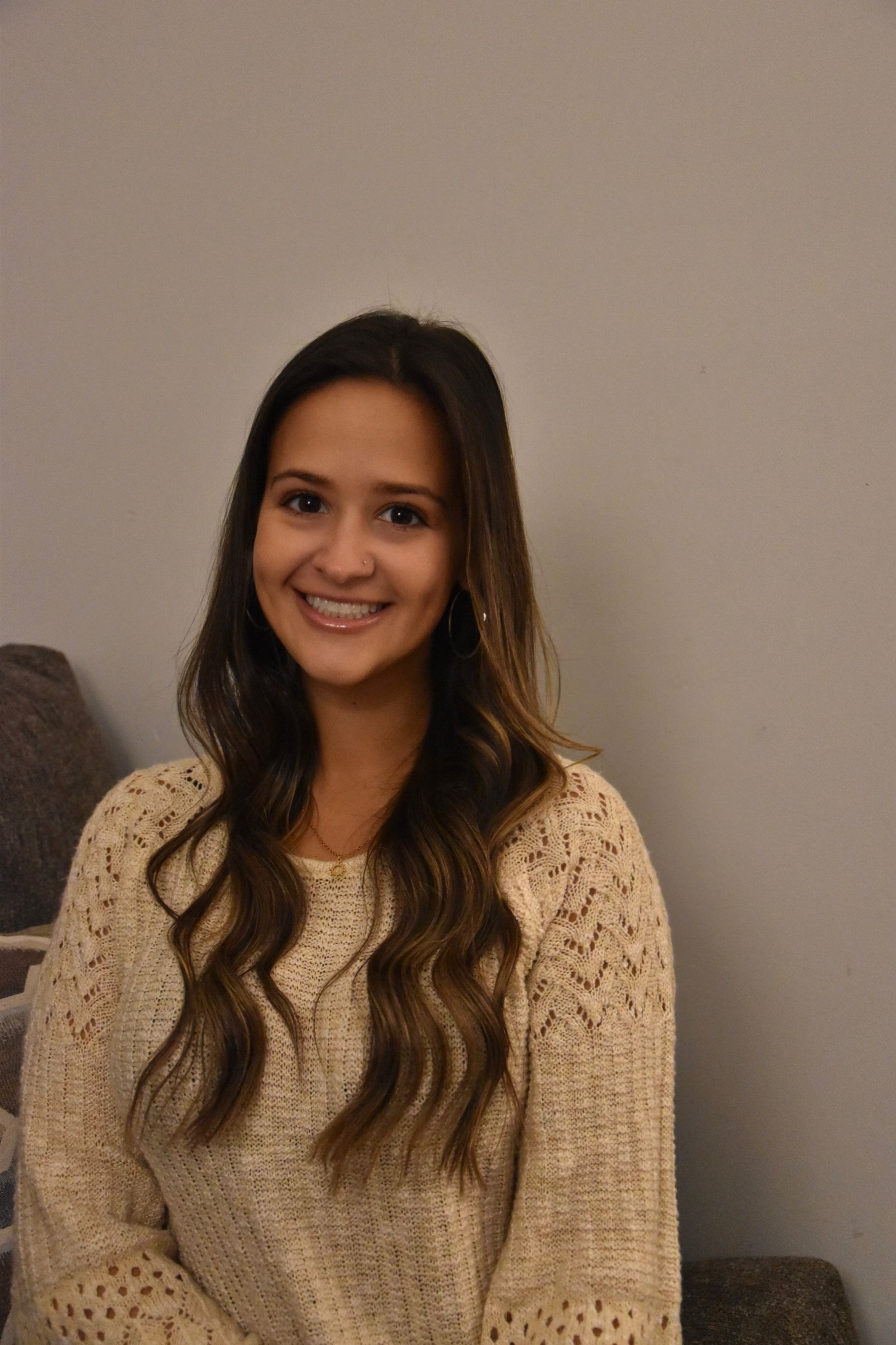 A smiling young woman with long, wavy brown hair wearing a beige crochet sweater, sitting on a sofa against a plain wall.