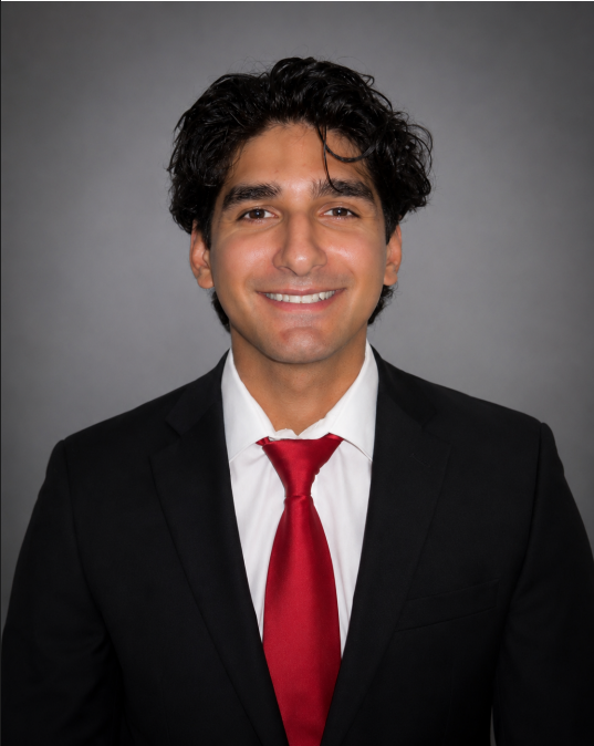 Headshot of a man in a black suit, white shirt, and red tie, smiling against a gray background.