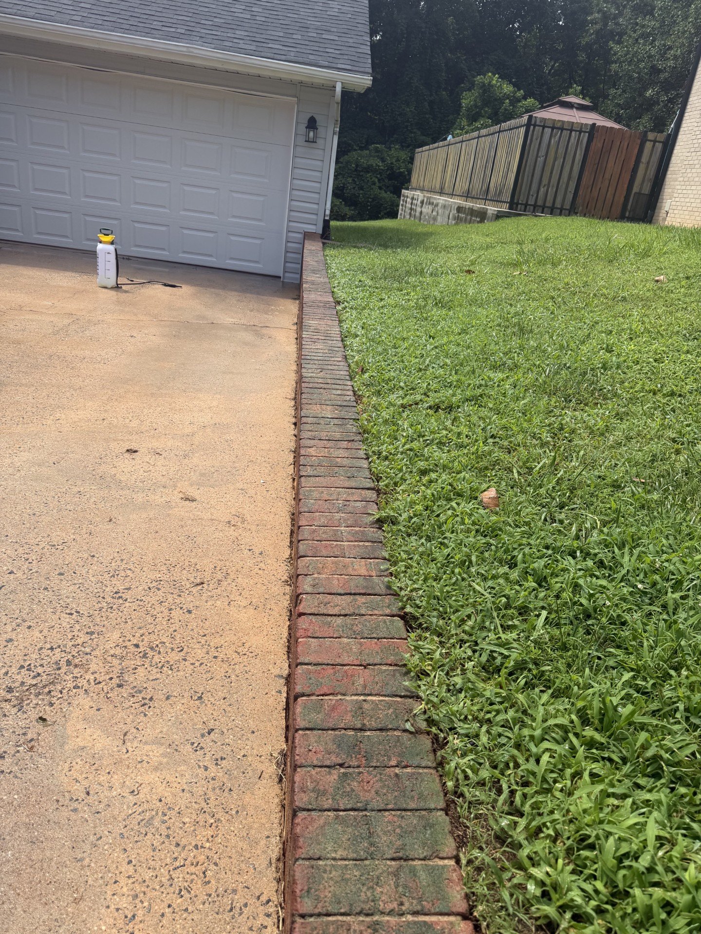 View of a sidewalk and a grassy yard with a brick border separating the sidewalk from the grass, with a house and a wooden fence in the background.