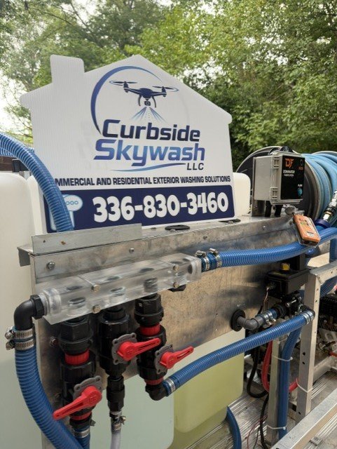Sign for Curbside Skywash LLC with phone number 336-830-3460, mounted on a machine with blue hoses and mechanical components, outdoors surrounded by green trees.