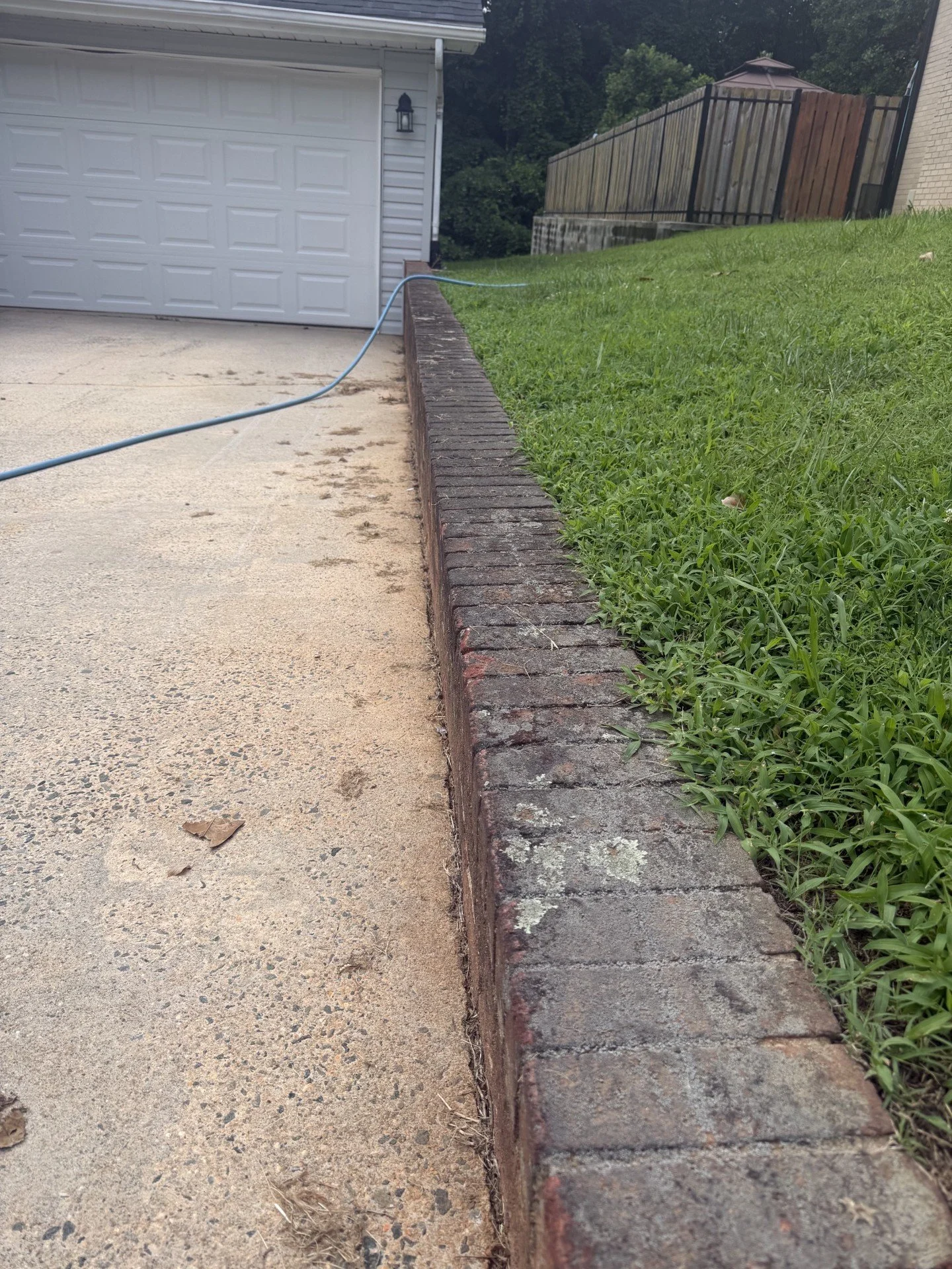 A concrete driveway with some dirt and debris, separated from a grassy yard by a brick border. There is a white garage door, a small wall-mounted light, and a wooden fence with trees in the background.