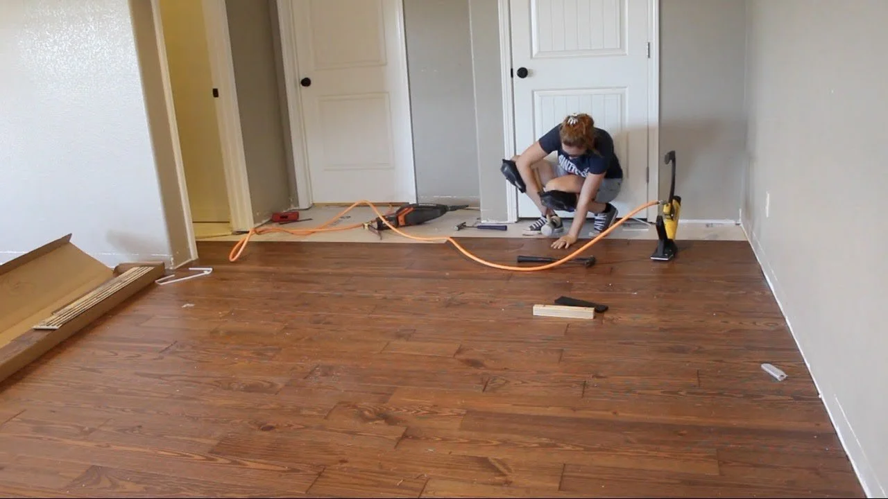 A woman installing or repairing hardwood flooring in a room, kneeling on the floor with tools and equipment around her.