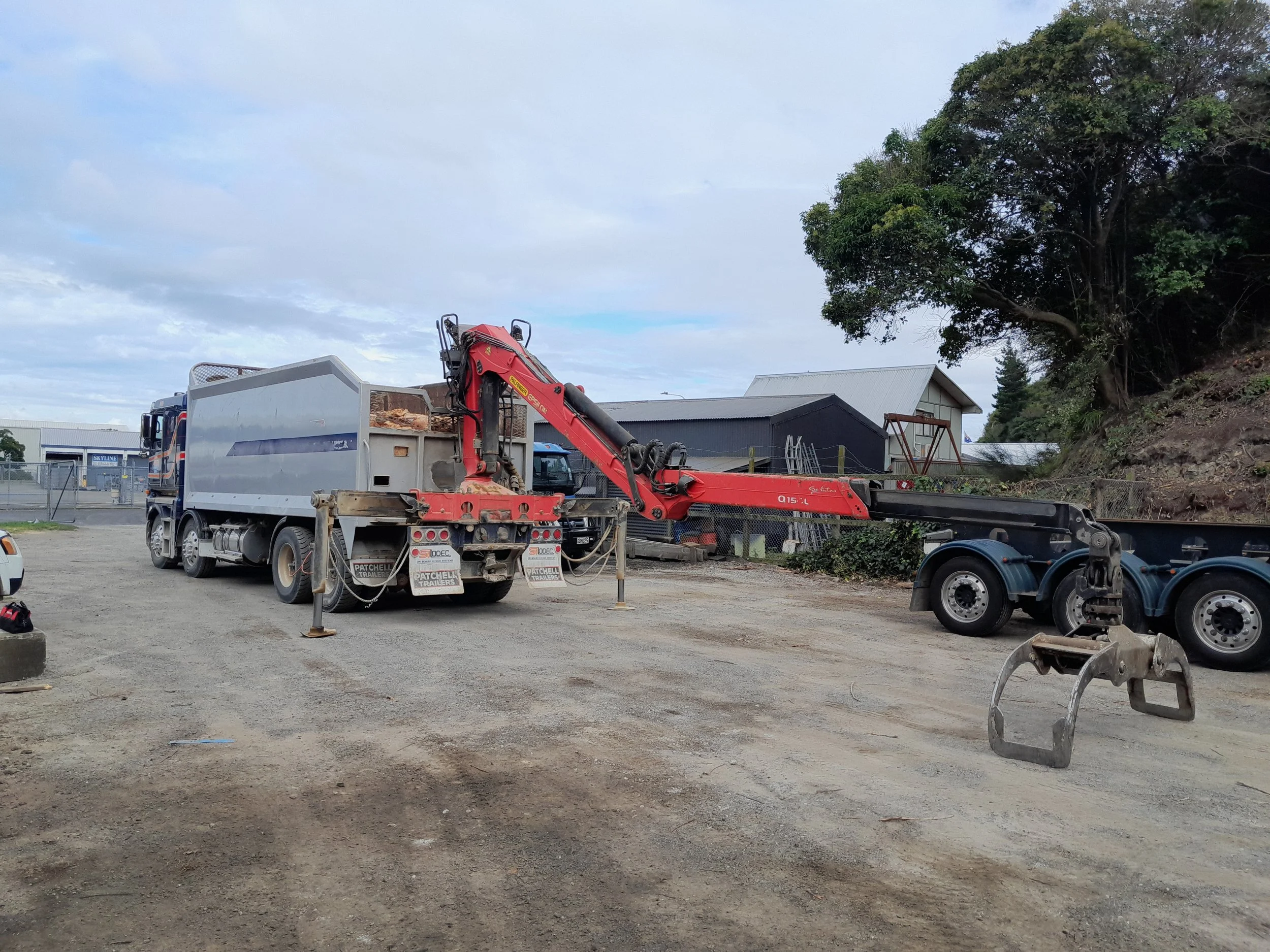 A construction site with a large white truck that has an attached red log loading handler, with graple. There are metal fences around the area, and there are some style buildings in the background, as well as a tree on the right side of the image.