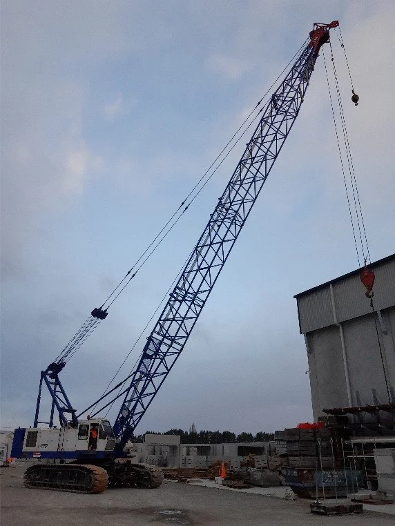 A large blue construction crane with a long boom extending upward at a construction site beaming over some buildings and equipment.