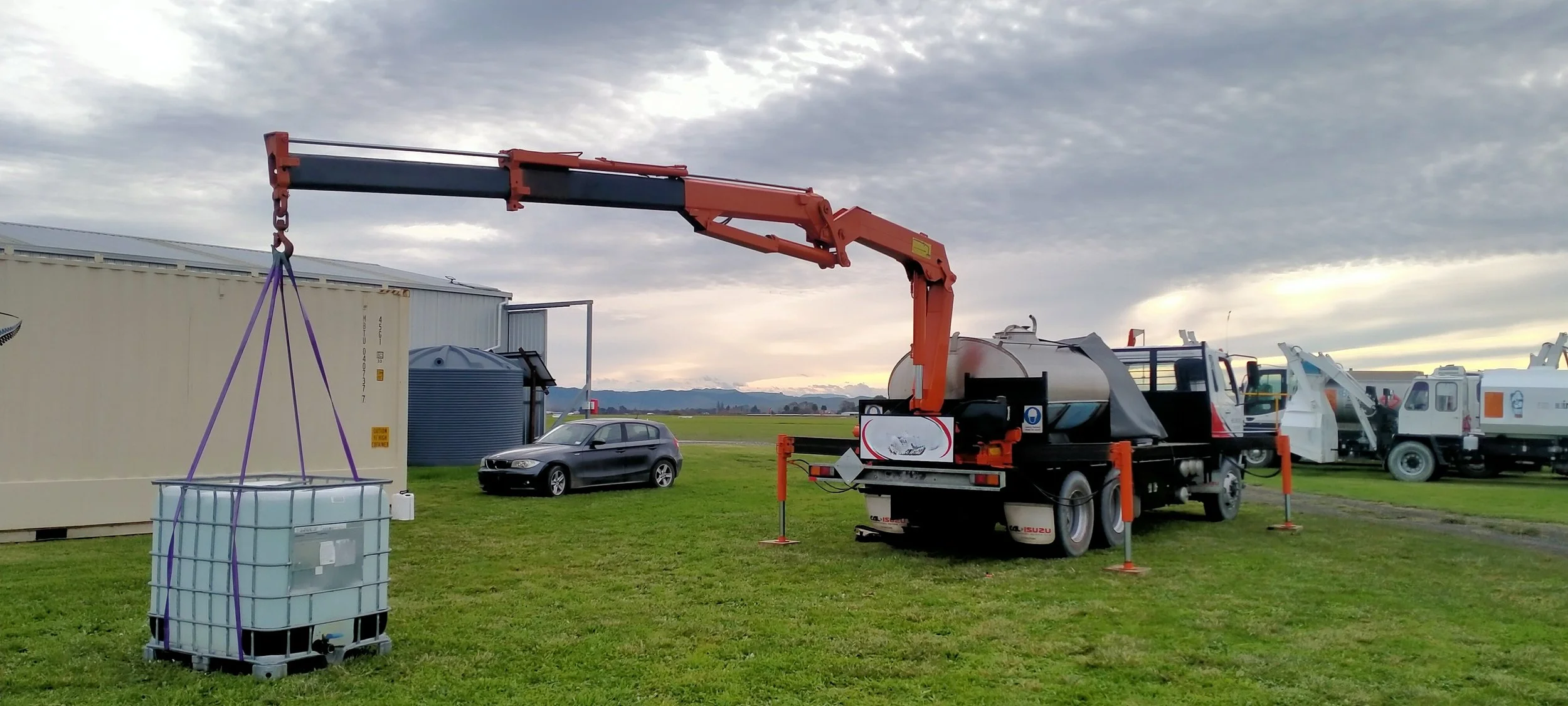 A crane truck lifting a large white water tank with purple straps in a grassy open area near a beige building and a parked gray car. Several other water trucks are lined up in the background, under a cloudy sky.