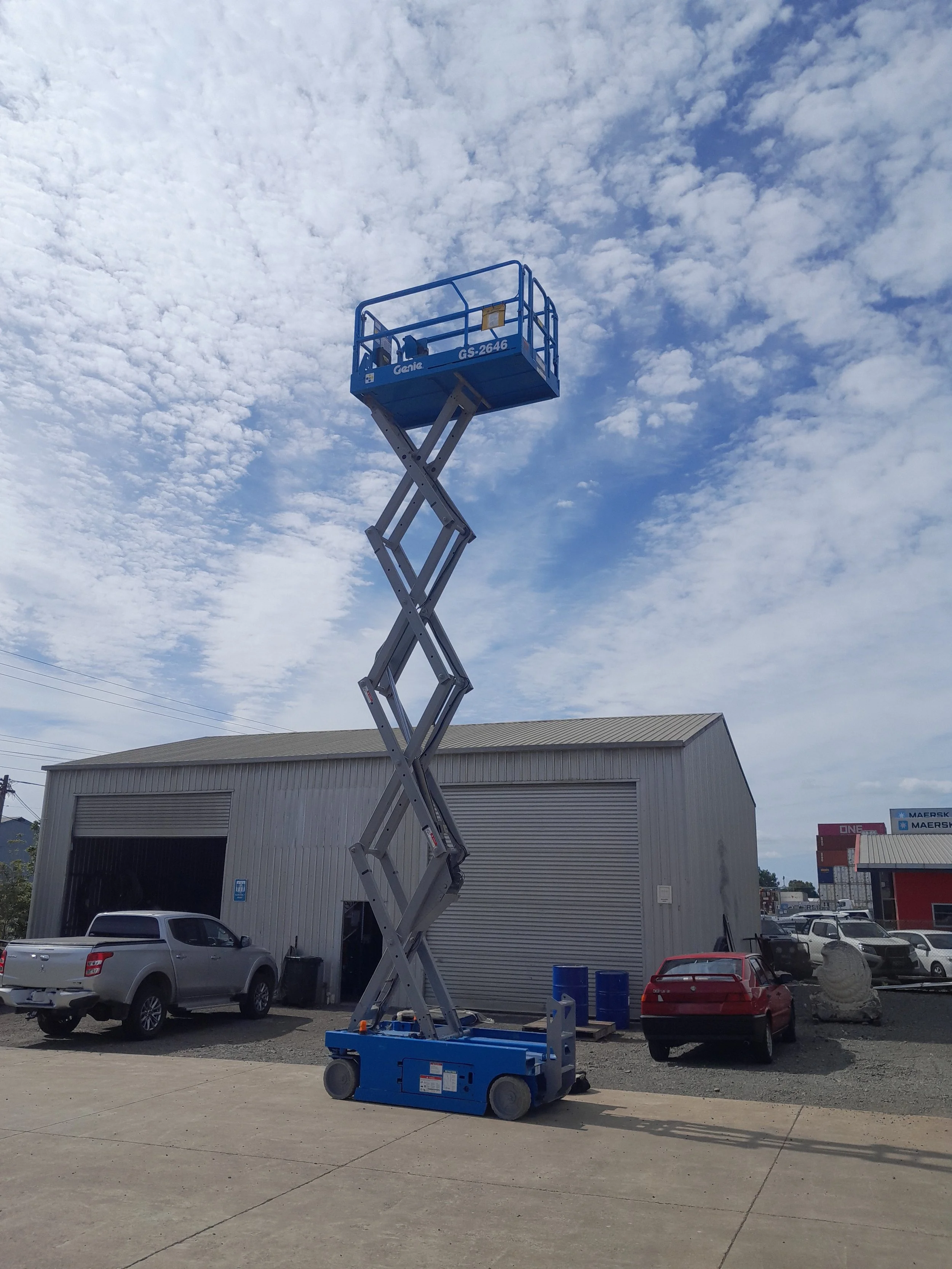 A blue scissor lift extended in an outdoor parking lot area near a large warehouse building. Several parked cars and shipping containers are visible in the background, with a cloudy sky overhead.