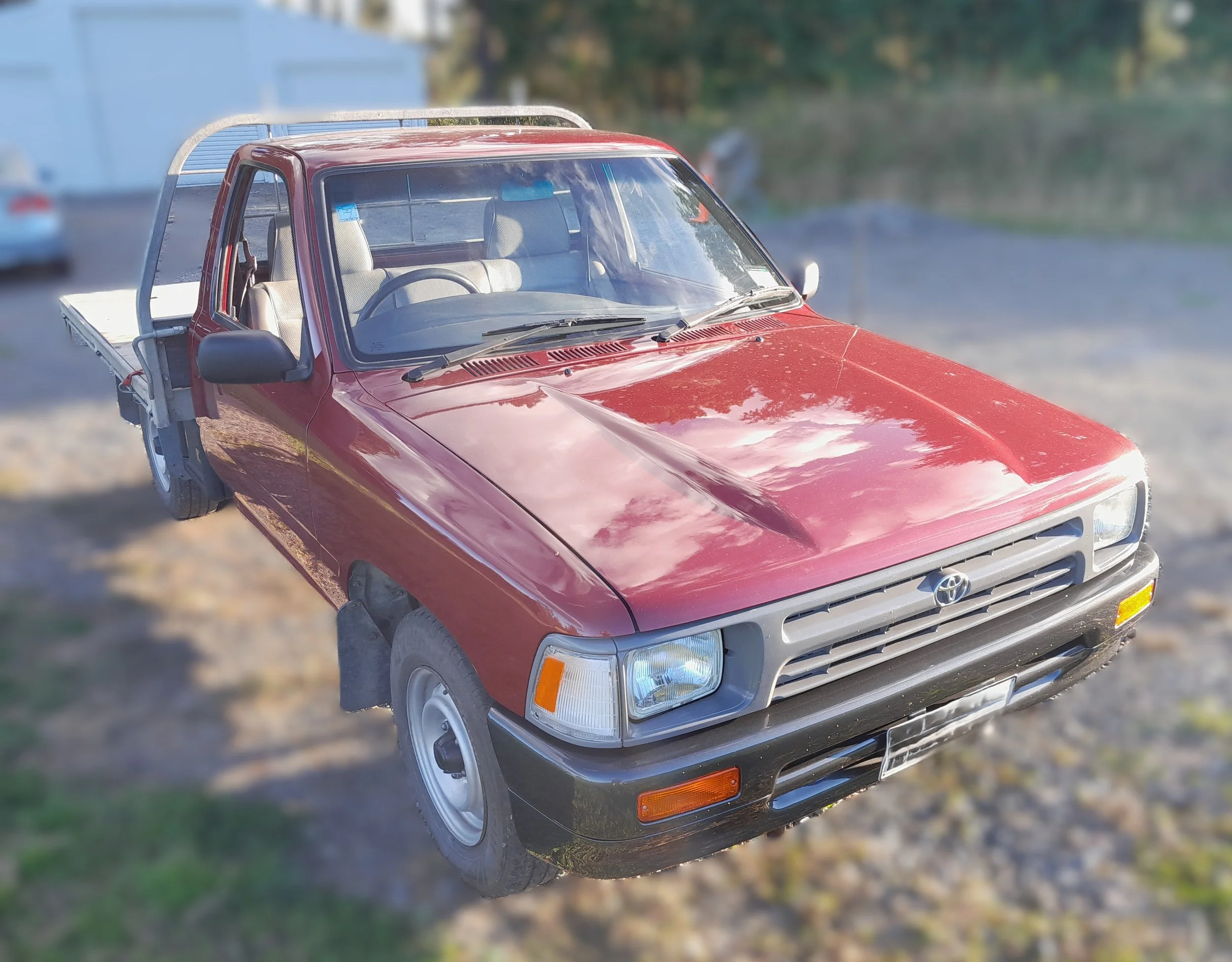 Close-up of a vintage red Toyota pickup truck parked outdoors, with a slightly blurred background of trees and grass.
