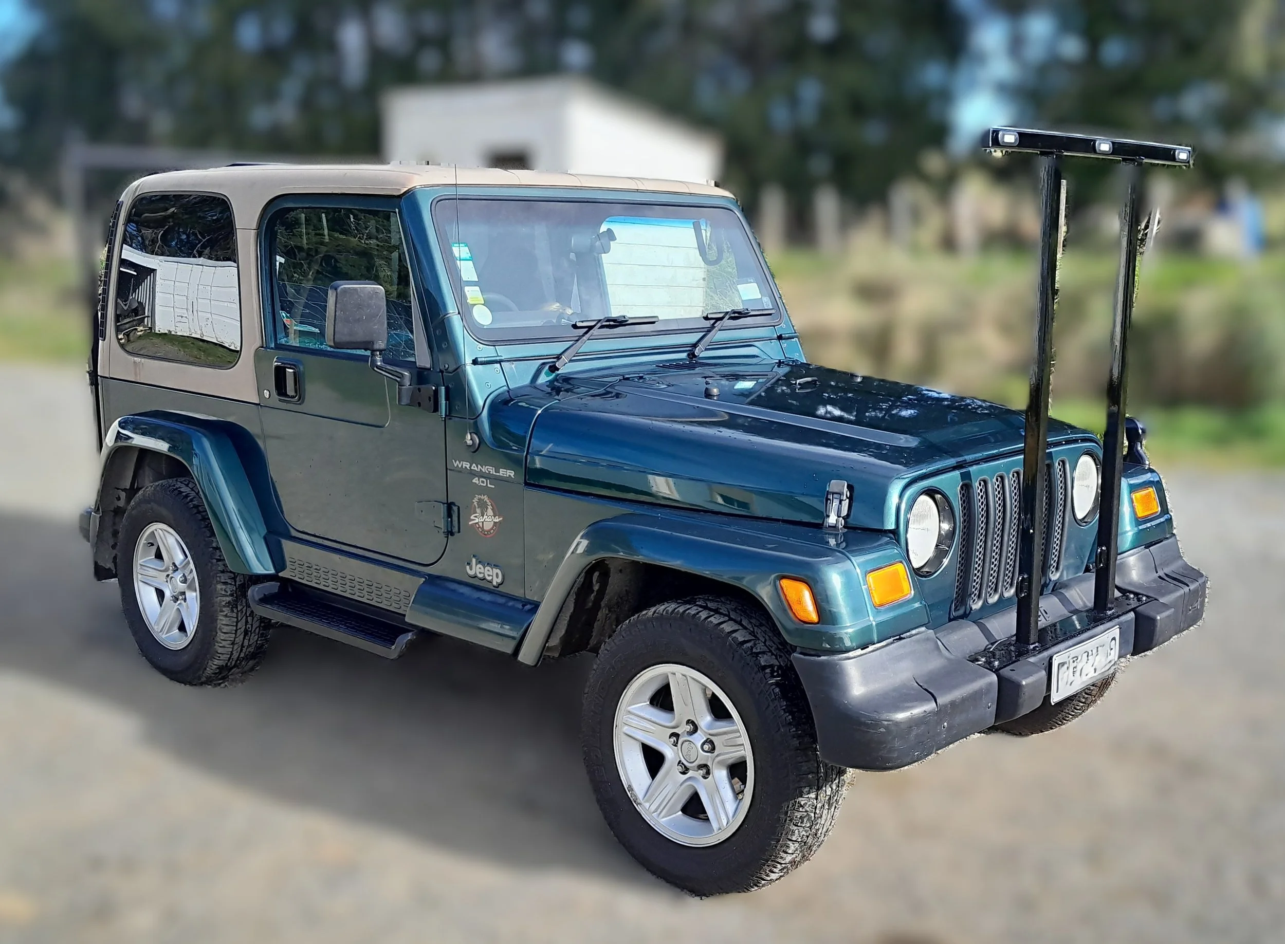 A blue Jeep Wrangler parked outdoors with two LED light bars mounted on the front bumper, reflected scenery on the windshield, and a mountainous background.