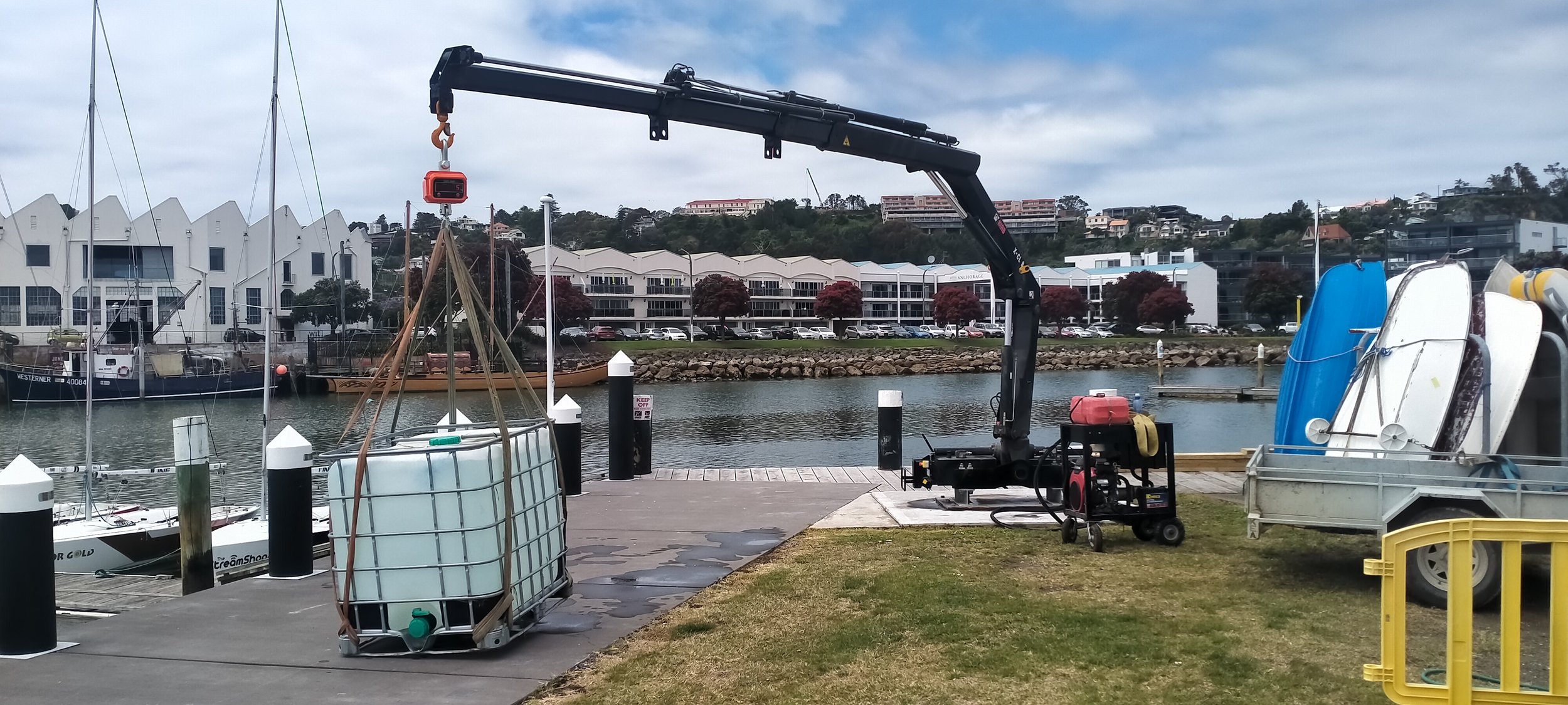 A marina with boats docked, a crane lifting a large container, and storage tanks on a dock, with buildings and hillside in the background.