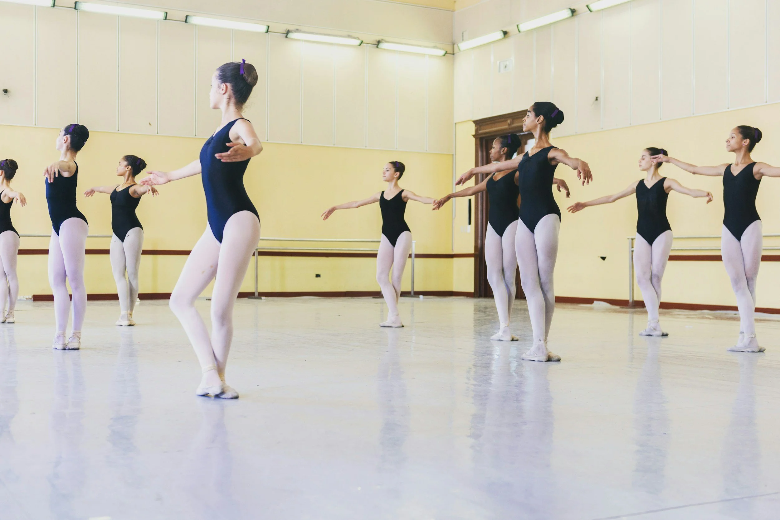 Group of ballet dancers practicing in a dance studio, wearing black leotards, white tights, and ballet shoes, with their hair tied back in buns, standing in a line with arms extended to the sides.
