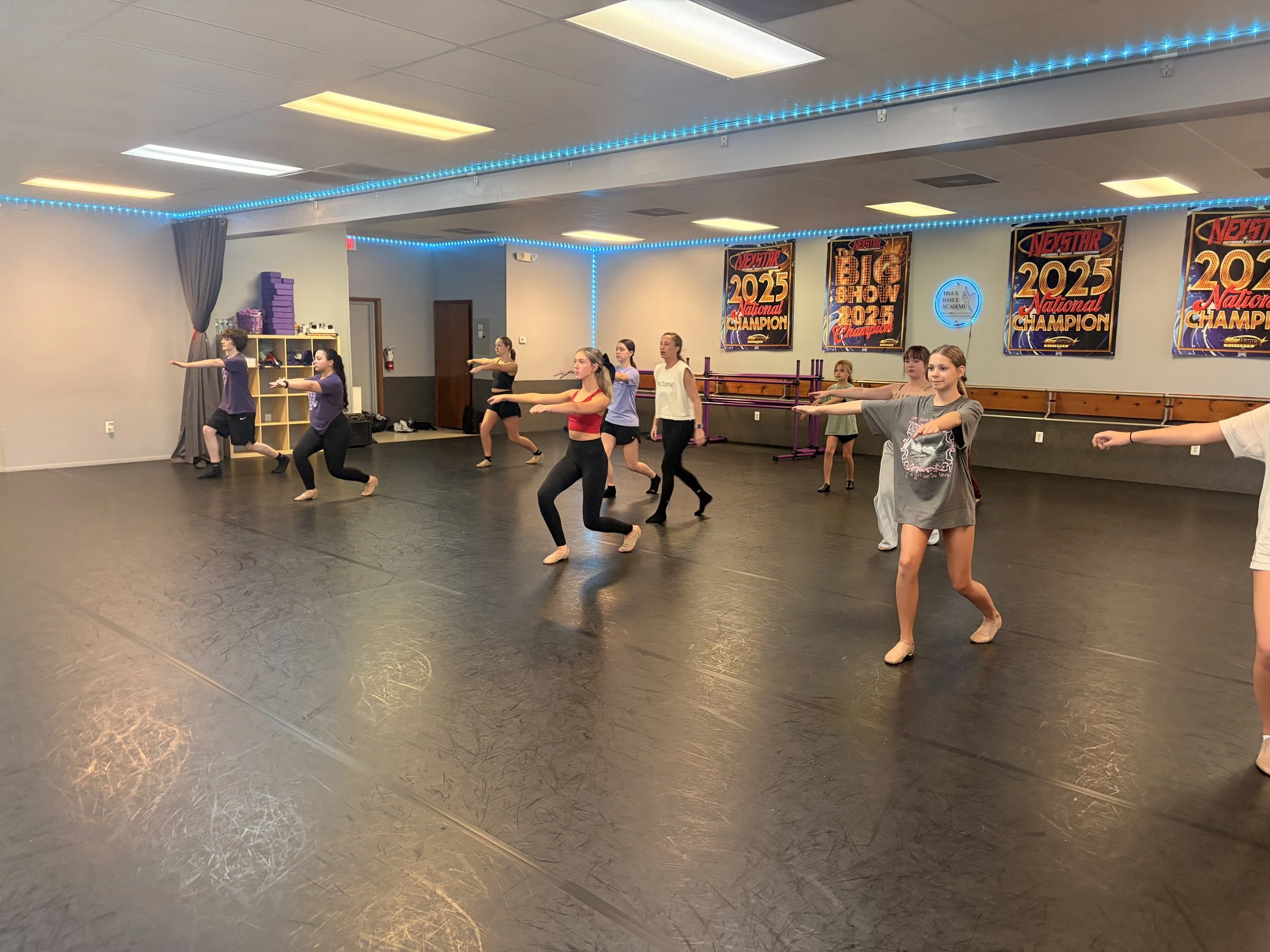 Group of young dancers practicing ballet in a dance studio with posters on the wall and blue LED lights along the ceiling.