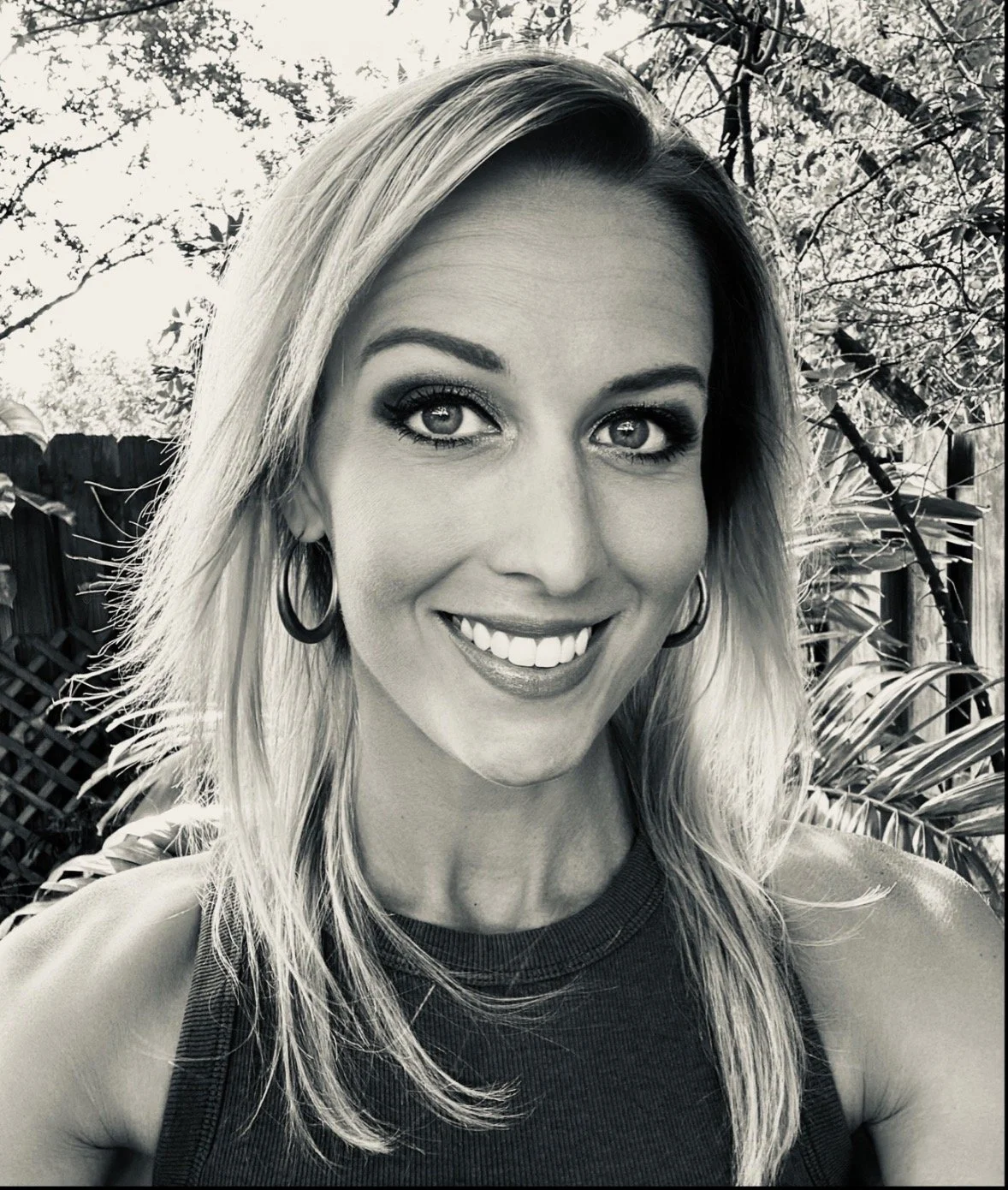 A smiling woman with shoulder-length blonde hair, wearing hoop earrings and a sleeveless top, standing outdoors with trees and plants in the background.