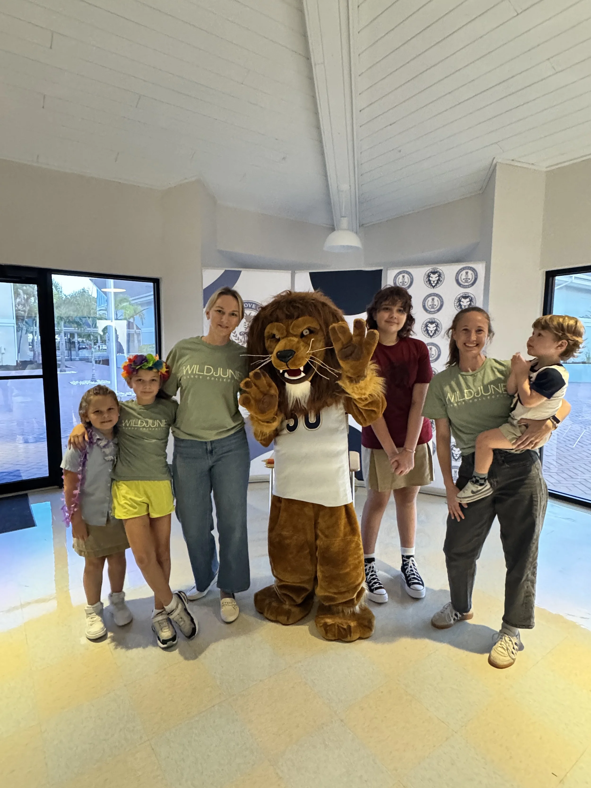 A group of children and adults standing with a person in a lion costume at an indoor location, with large windows and a backdrop featuring lion emblems.