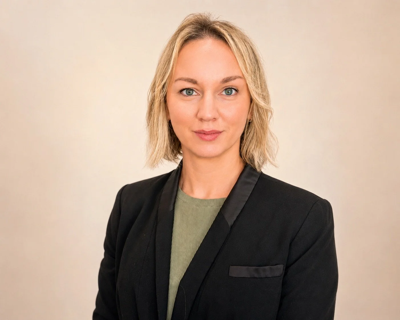 A woman with shoulder-length blond hair, blue eyes, wearing a black blazer over a green top, standing against a plain beige background.