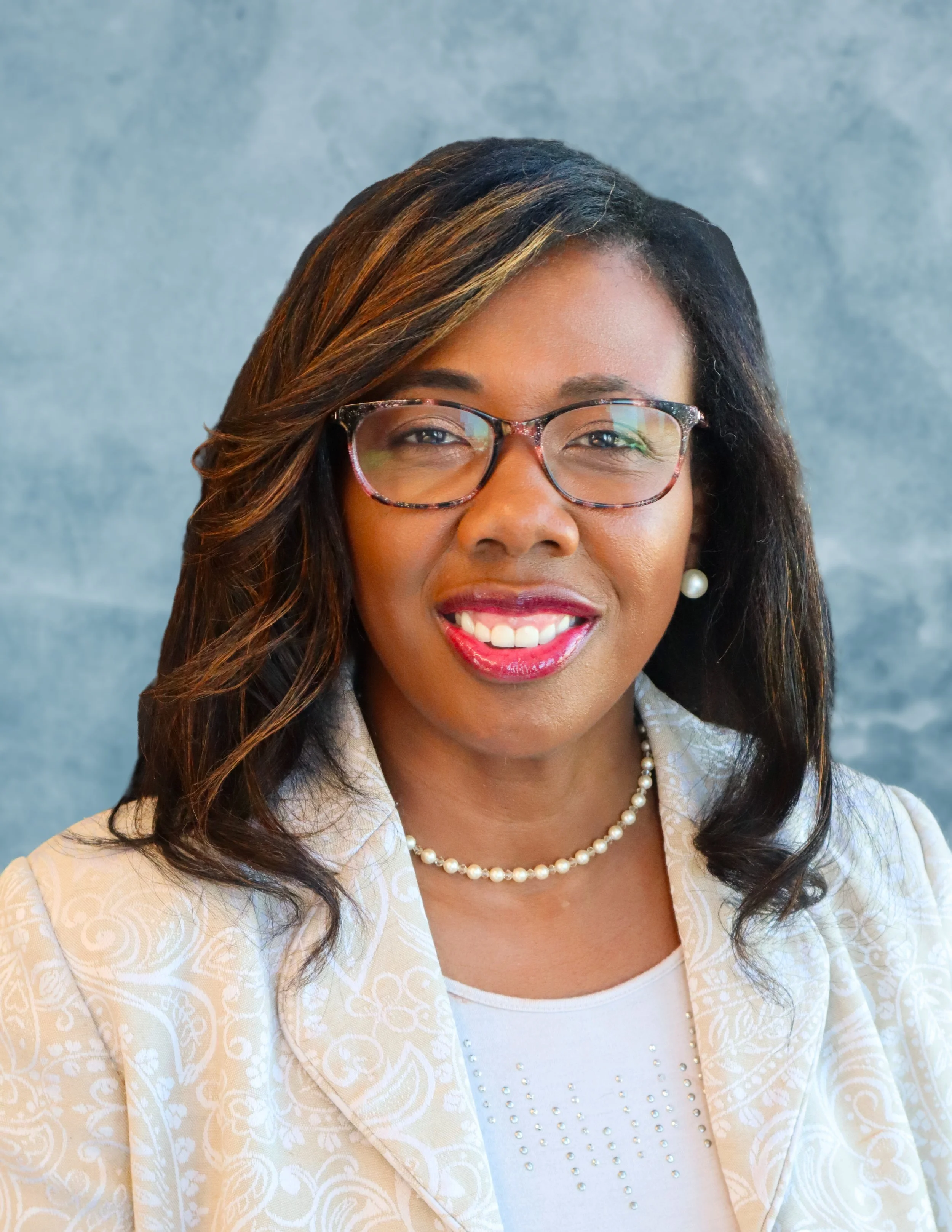 Close-up of a smiling woman with glasses, pearl earrings, and a cream-colored patterned jacket against a blue background.
