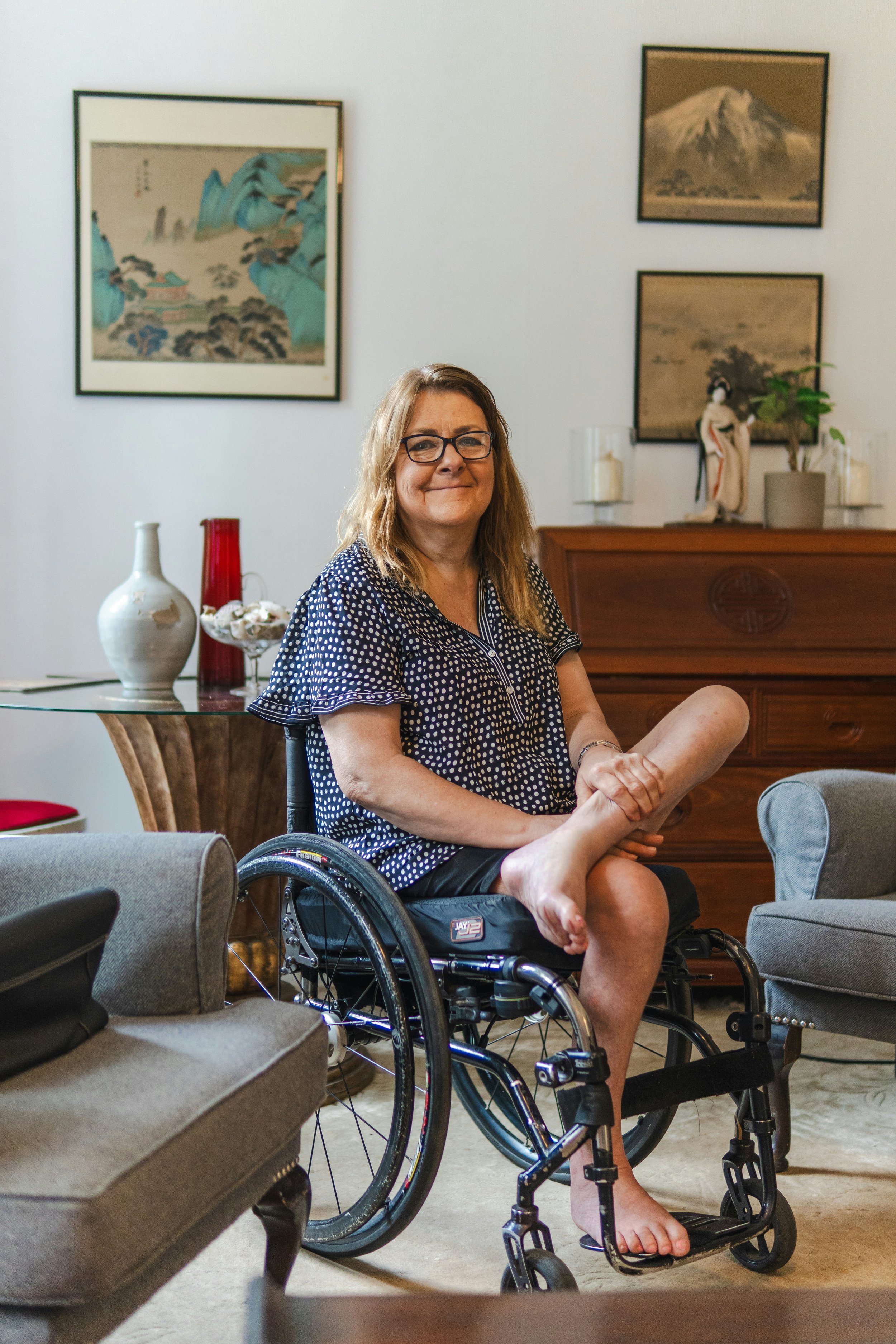 A woman with glasses in a polka-dotted shirt sitting in a wheelchair inside a living room, smiling at the camera. The room is decorated with framed artwork, vases, and a wooden dresser with a small plant.
