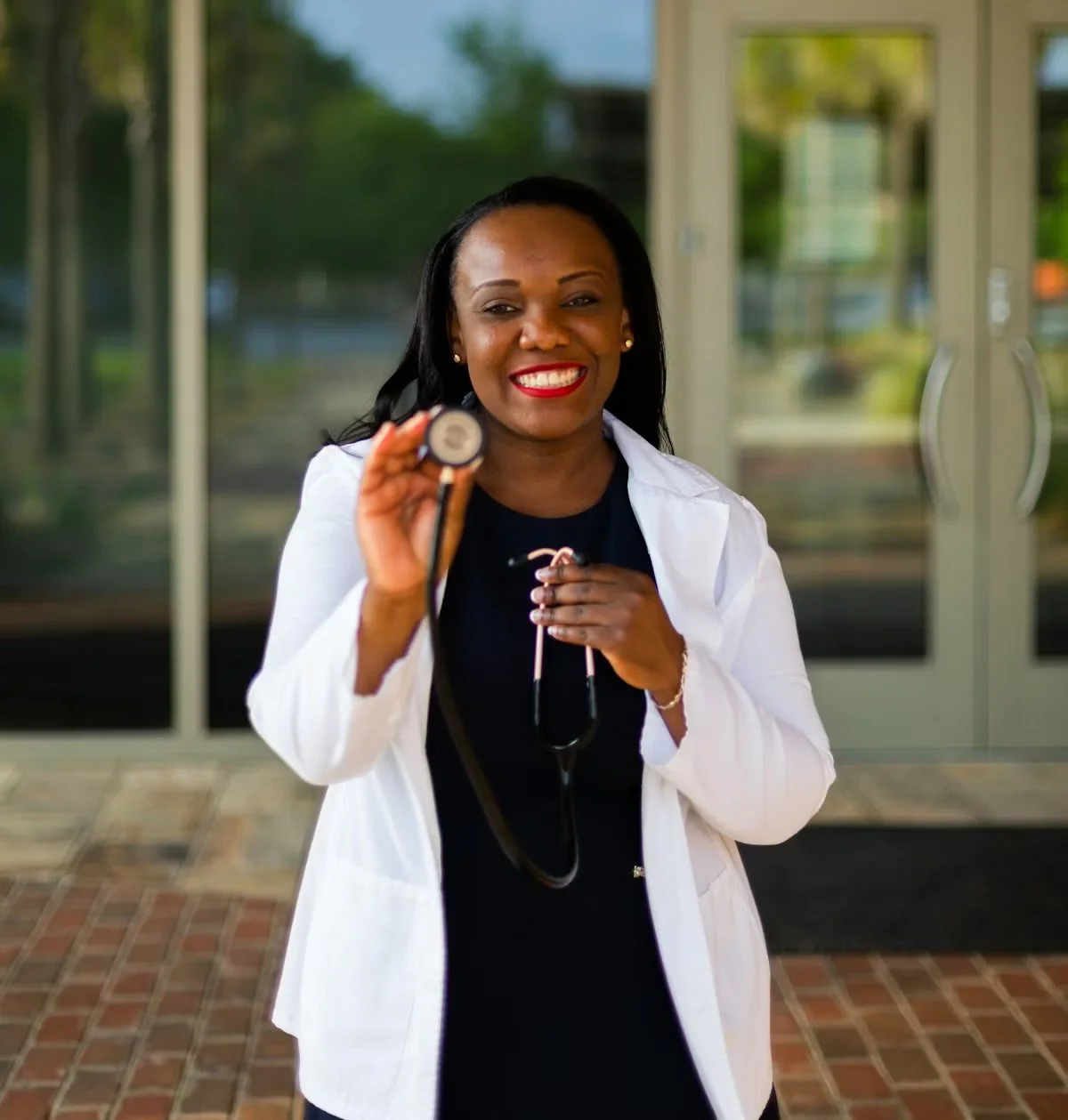 A smiling female doctor or healthcare professional holding a stethoscope outside a medical facility.