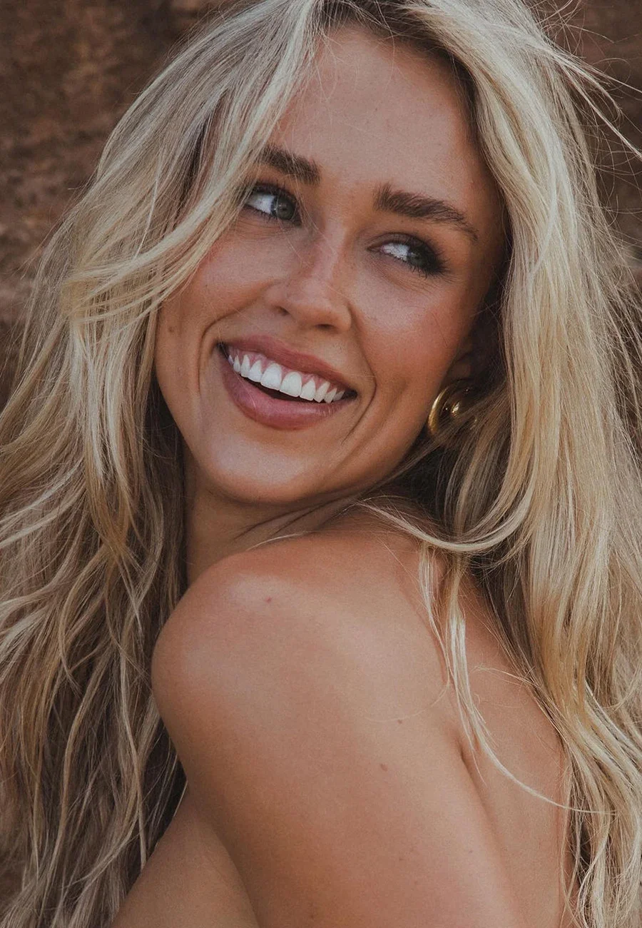 Zoe Creed with long blonde hair smiling with white teeth, looking to the side, showing her shoulder and part of her back.