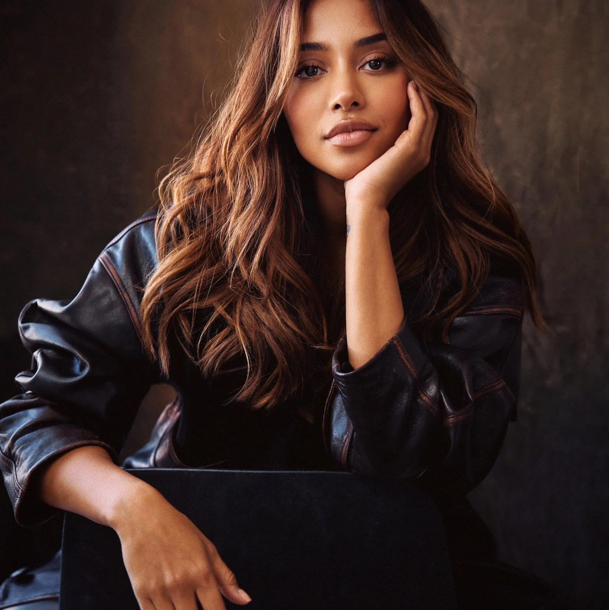 Maria Thattil  with long, wavy brown hair resting her face on her hand, wearing a black leather jacket, against a dark background. With Australian pageant walk coach Thelma.