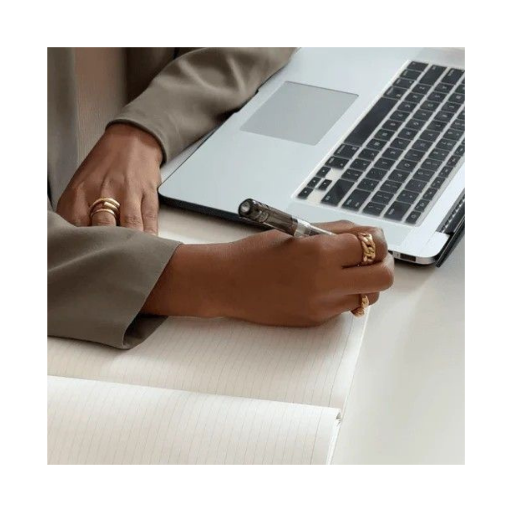 Person writing in a notebook with a laptop nearby, wearing rings on their fingers.