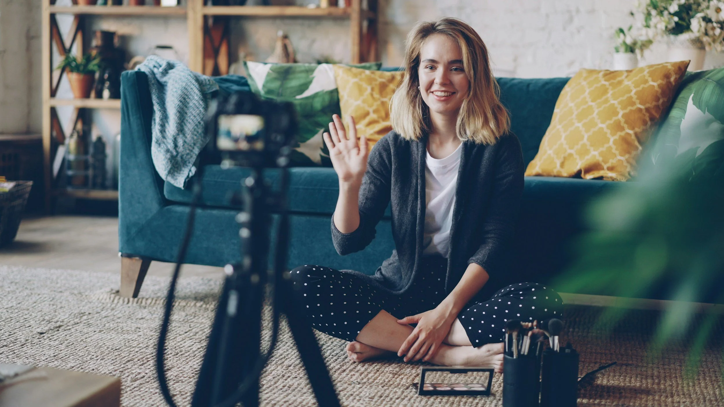 A young woman with shoulder-length blonde hair, wearing a dark cardigan and polka-dot pants, sitting cross-legged on a rug in a living room, smiling and waving at a camera. The room has colorful pillows on a teal sofa, a wooden bookshelf with plants and decorative items, and a textured white wall.