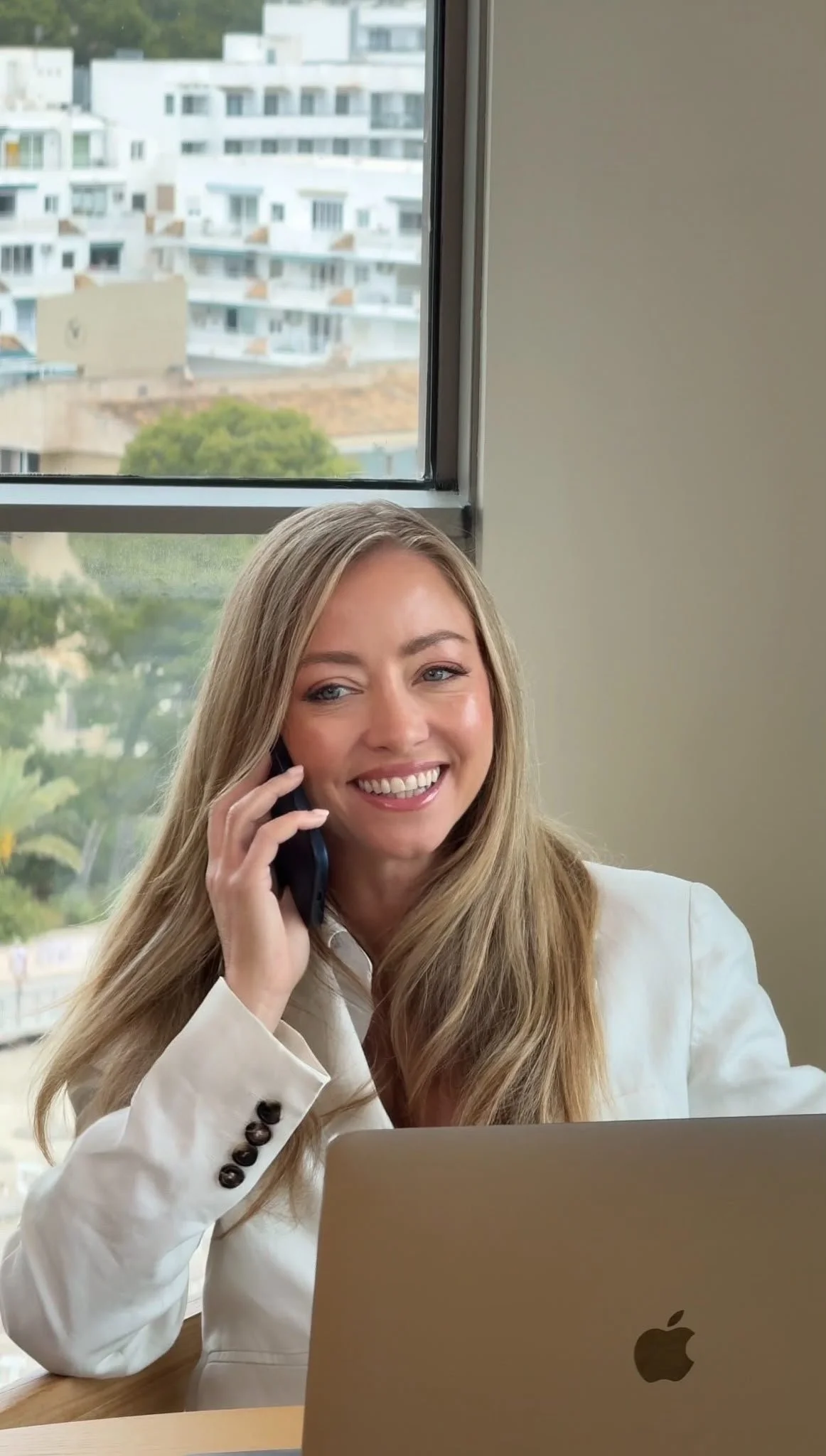 A woman with long blonde hair, wearing a white blazer, is smiling and talking on a phone at a desk with a laptop. There is a large window behind her showing a view of modern white apartment buildings and some trees.