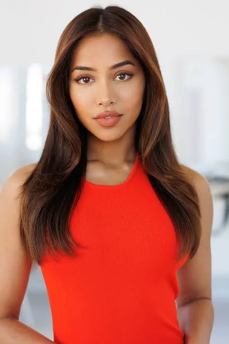 A young woman with long brown hair wearing a red sleeveless top standing indoors.