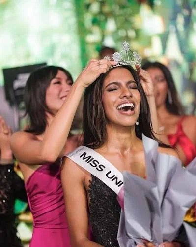 A woman wearing a sash that says 'Miss Uni' and a tiara is being crowned at a beauty pageant. She is smiling and surrounded by others celebrating.
