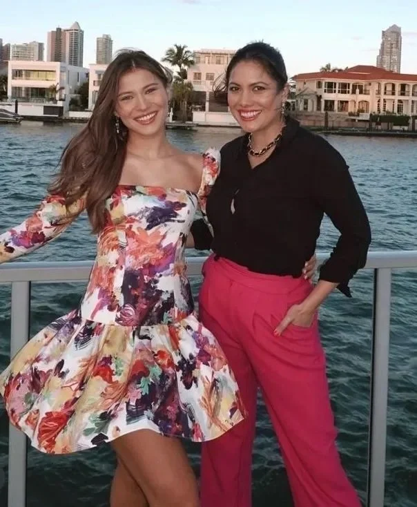 Two women smiling and posing on a boat by the water with city buildings in the background. One woman is wearing a colorful floral dress, and the other is wearing a black top with pink pants.
