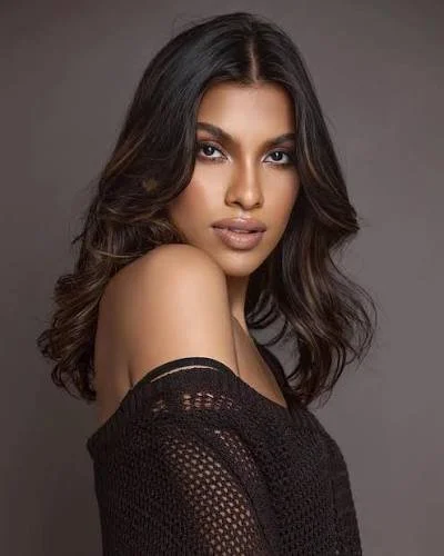 Priyanka Annuncia with long, wavy dark hair, wearing a black off-the-shoulder sweater against a gray background.