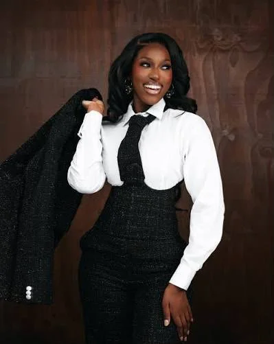 A woman with black hair and dark skin, wearing a white blouse, black tie, and black skirt, holding a black jacket over her shoulder, standing in front of a wooden background, smiling.