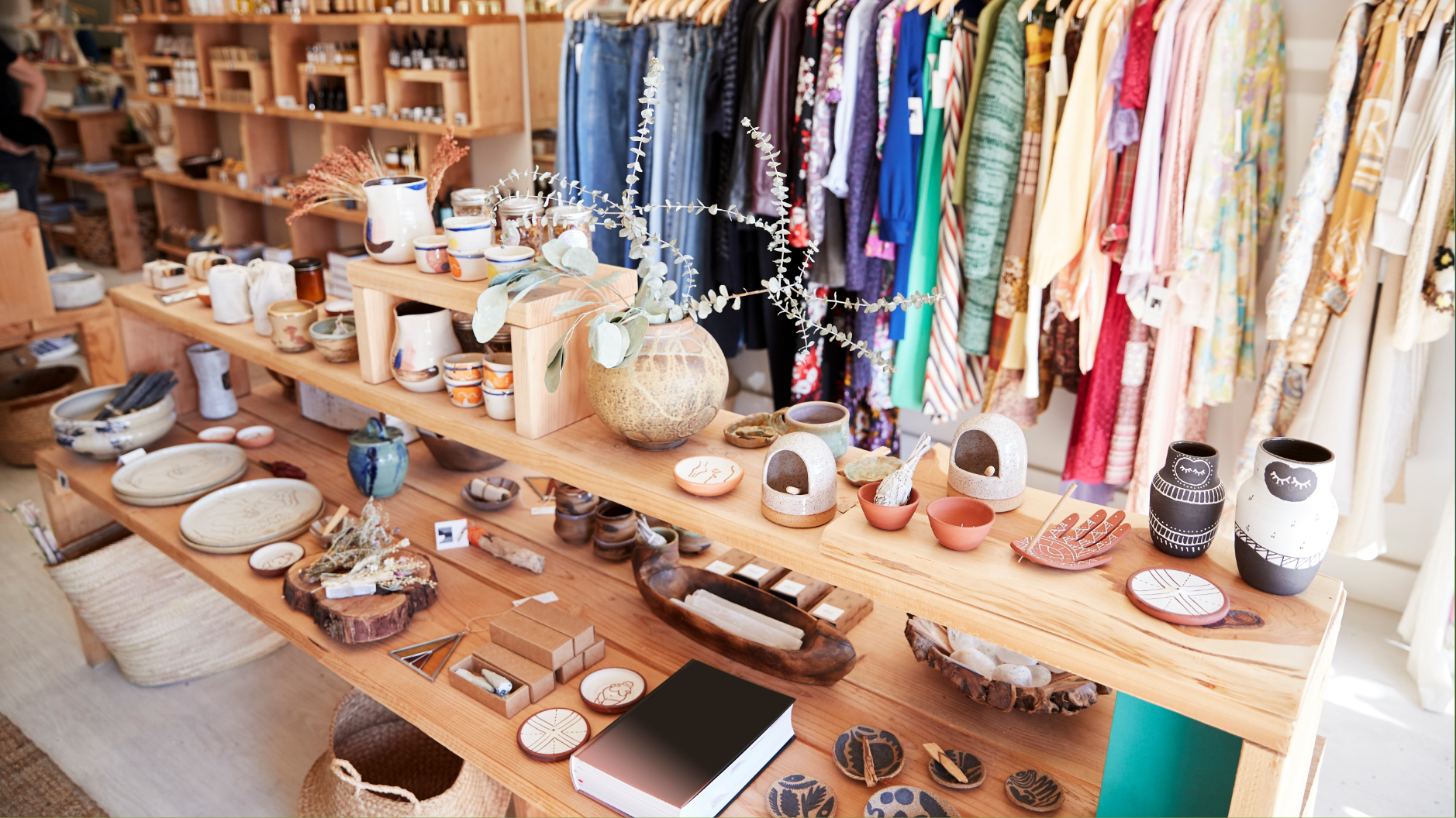 Display of various handmade pottery and ceramics on a wooden table in a boutique shop, with clothing racks in the background.
