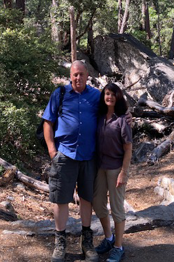 Pamela West and her spouse standing outdoors in Yosemite with large rocks and trees in the background, smiling.