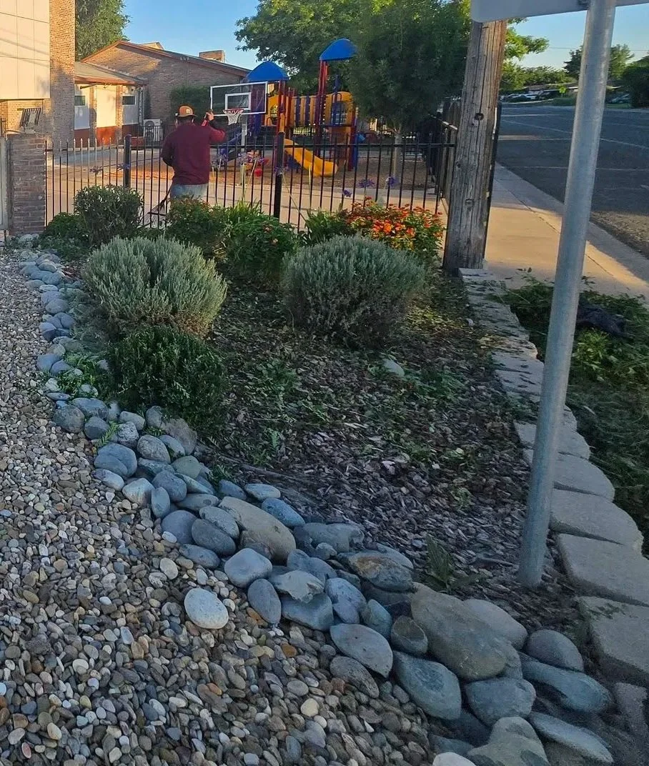 A person standing near a fenced playground with a colorful slide inside a residential area. The person, seen from behind, is wearing a dark jacket and a cap. The foreground features decorative rocks and small bushes, with a sidewalk and street on the right side.