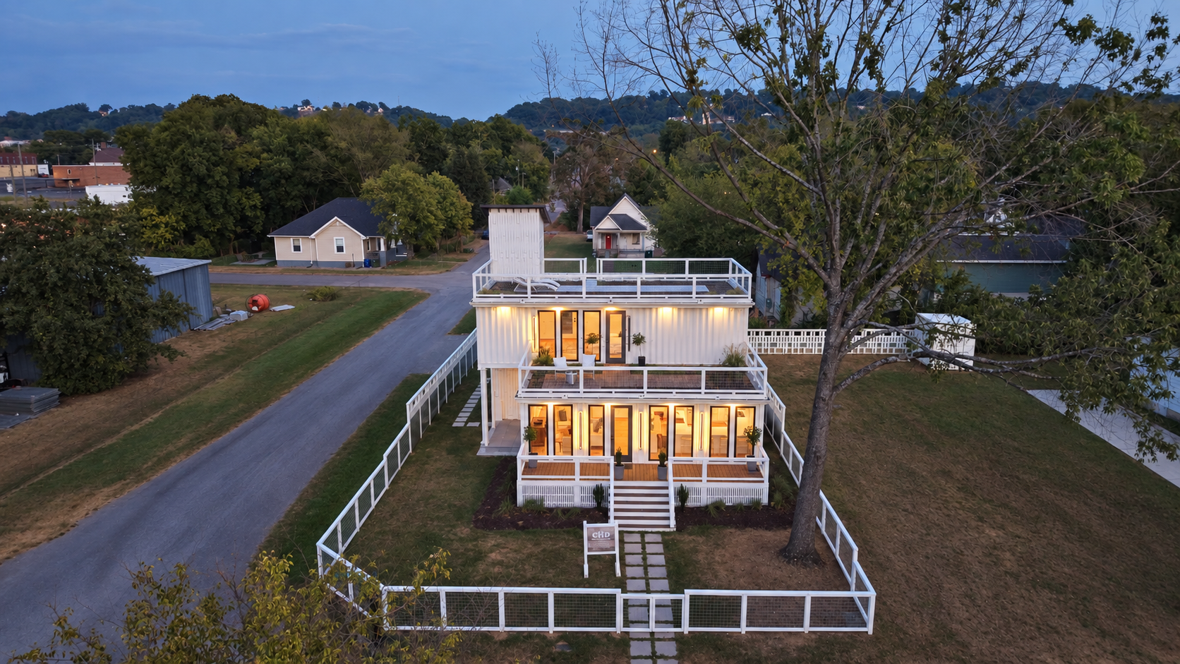 A modern, multi-story house with large glass doors and balconies, brightly lit from inside, surrounded by a white fence, with a yard and a large tree in front, at dusk.