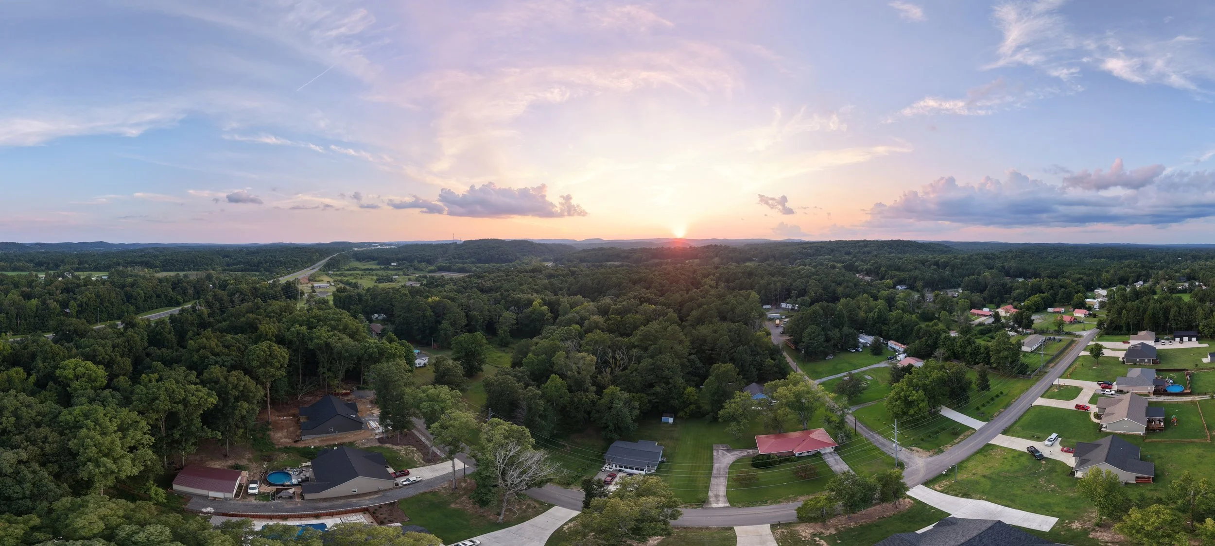 Aerial view of a suburban neighborhood with houses, trees, and roads at sunset, with a partially cloudy sky.