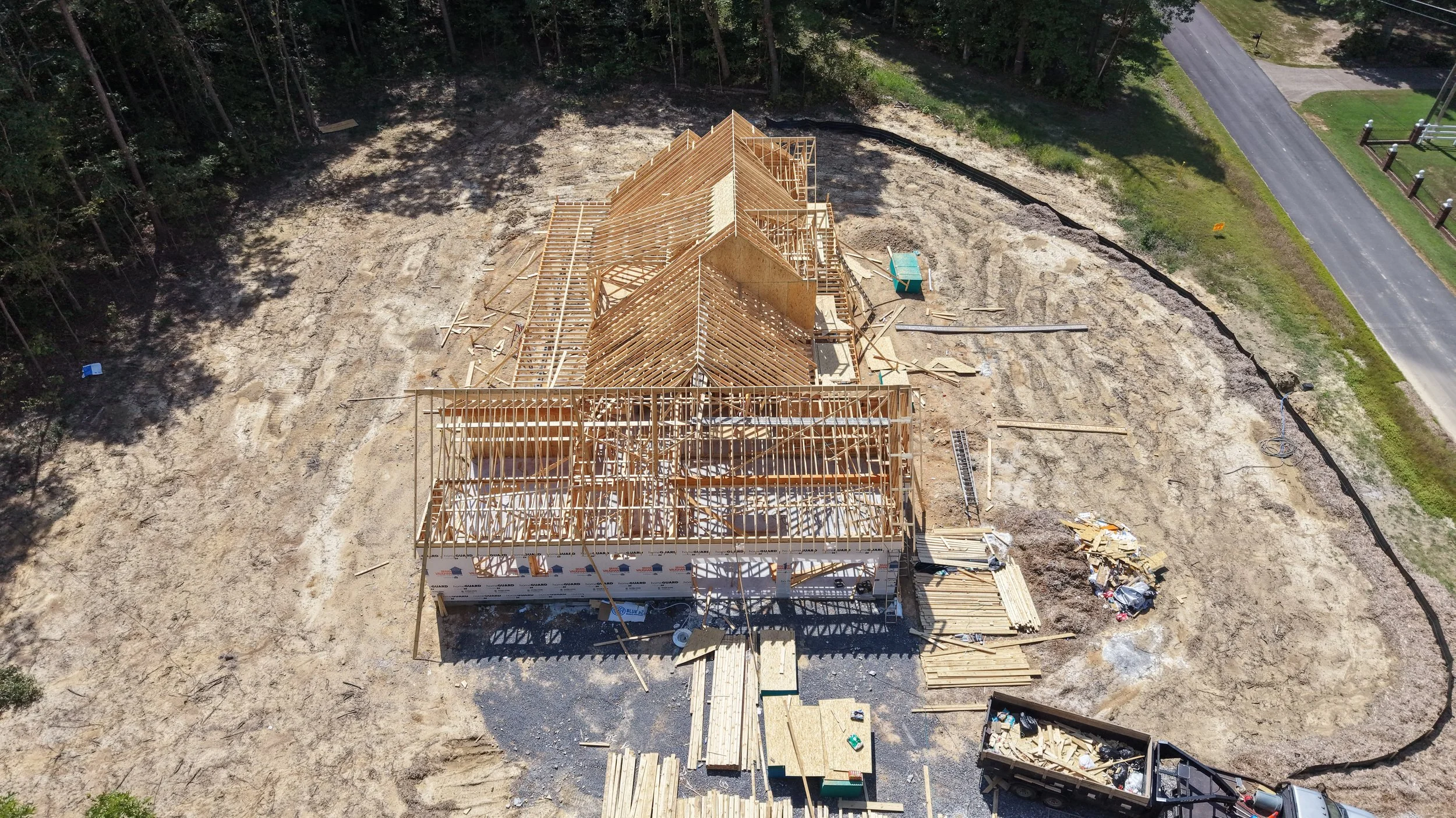 An aerial view of a house under construction with a wooden framework, situated on a cleared dirt lot surrounded by trees and roads.