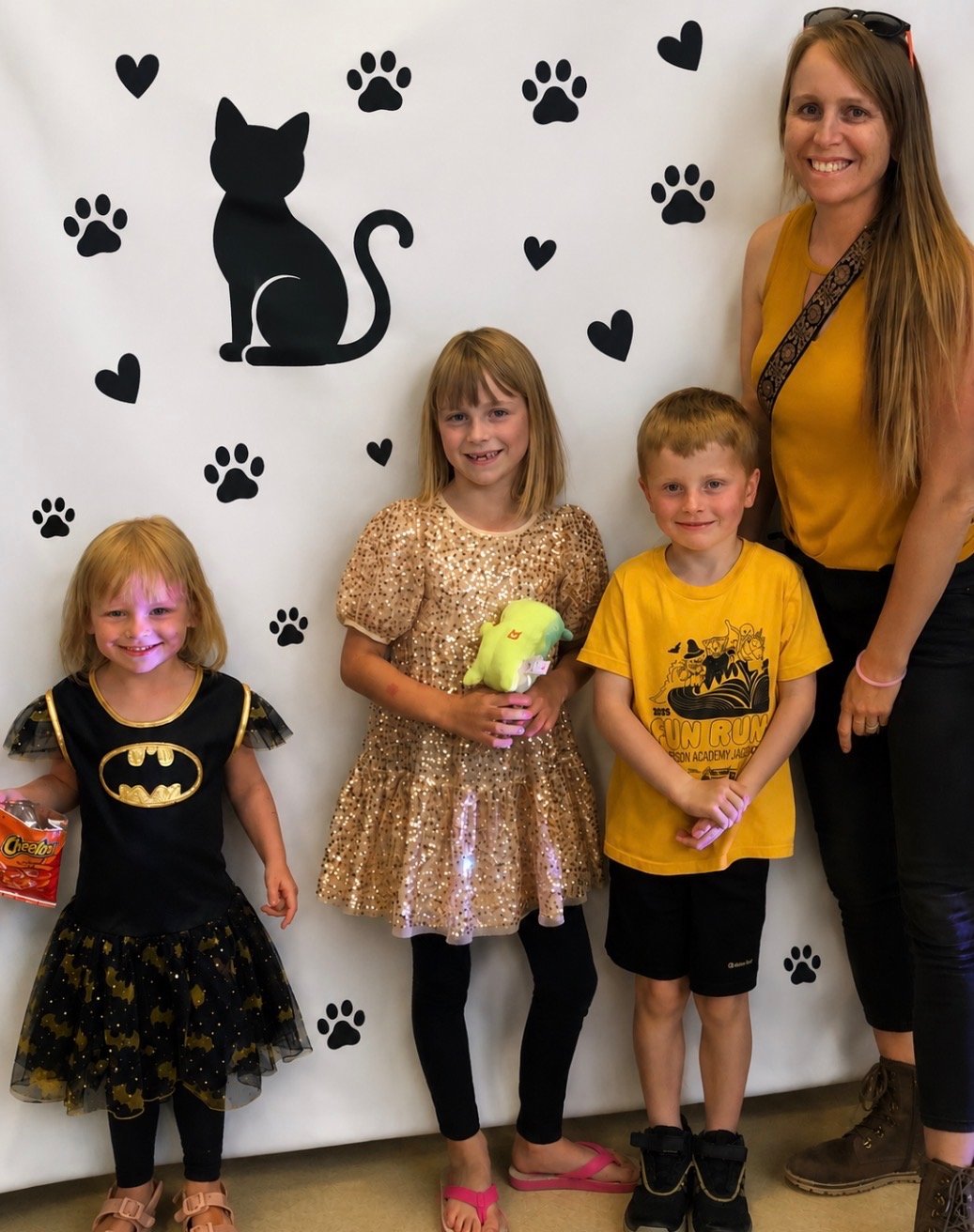 Four children and one adult woman standing in front of a white wall decorated with black silhouette images of a sitting cat, paw prints, and hearts. The children are dressed in costumes or casual clothing, and they are smiling and holding small plush toys or snacks.
