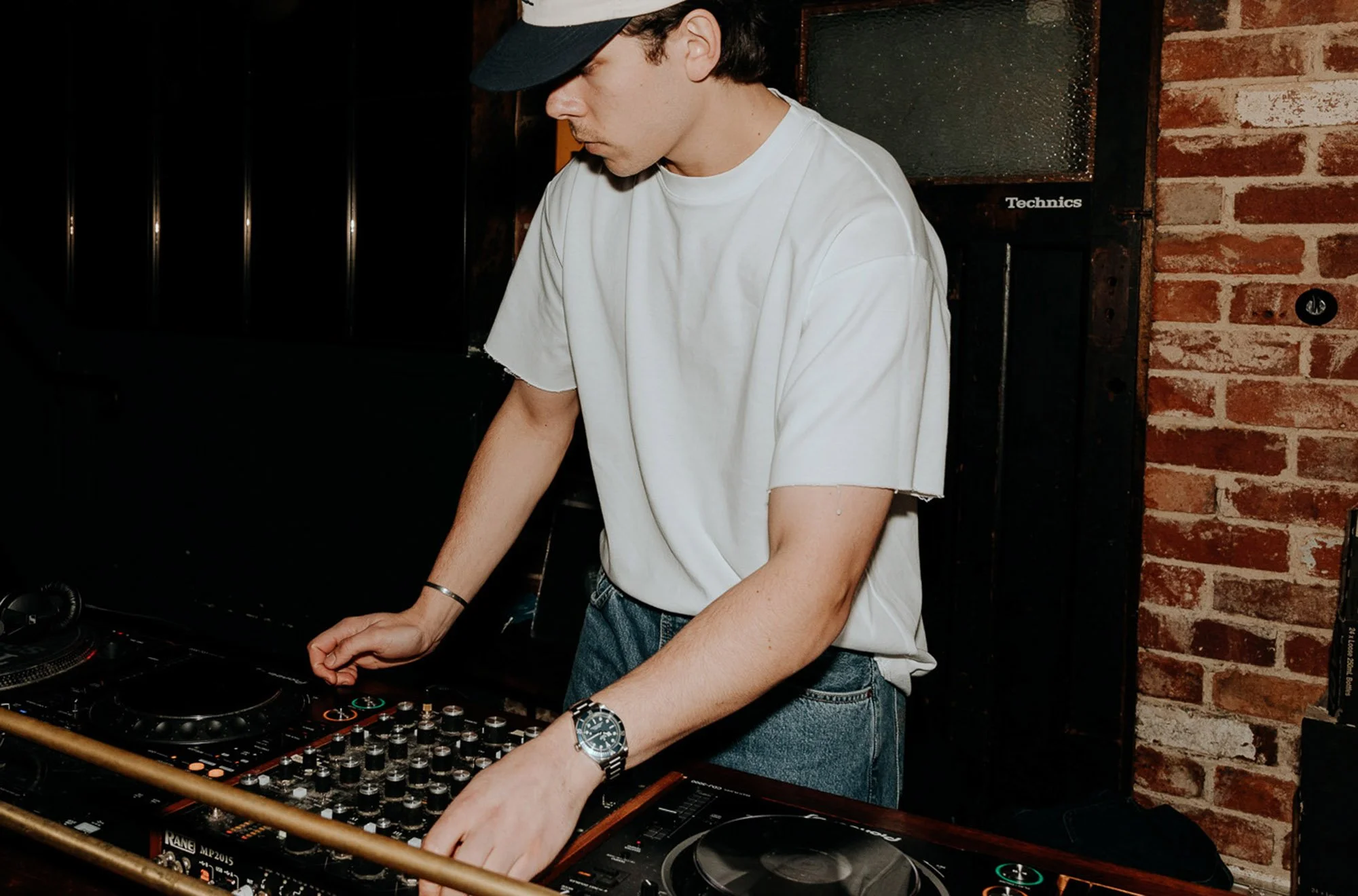 A young man in a white T-shirt, blue jeans, and a black cap, wearing a wristwatch, is DJing with mixer and turntables in a dimly lit music venue with brick and wood walls.