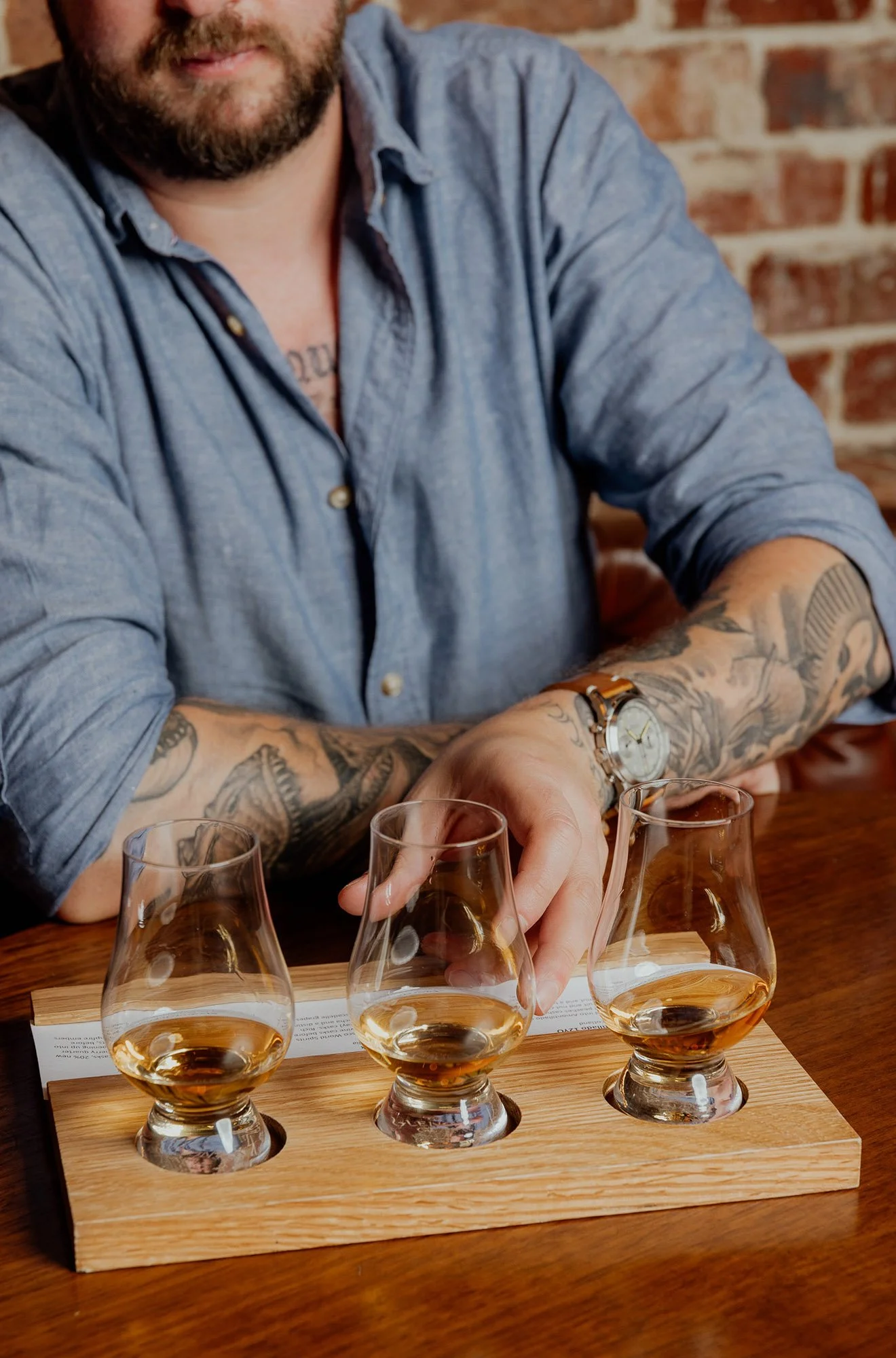 A man with tattoos on his arms and a beard wearing a blue shirt, seated at a table with a whiskey tasting flight, holding a glass. The flight has five glasses filled with whiskey, and there is a tasting note sheet beneath the glasses. The background features a brick wall.