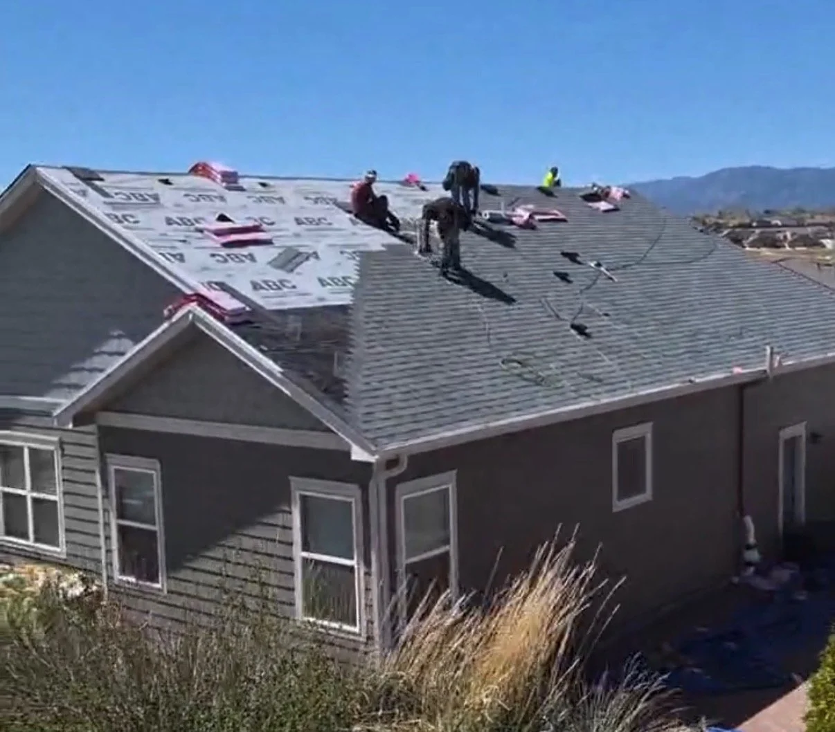 Workers installing roof shingles on a house with a gray exterior in sunny weather.