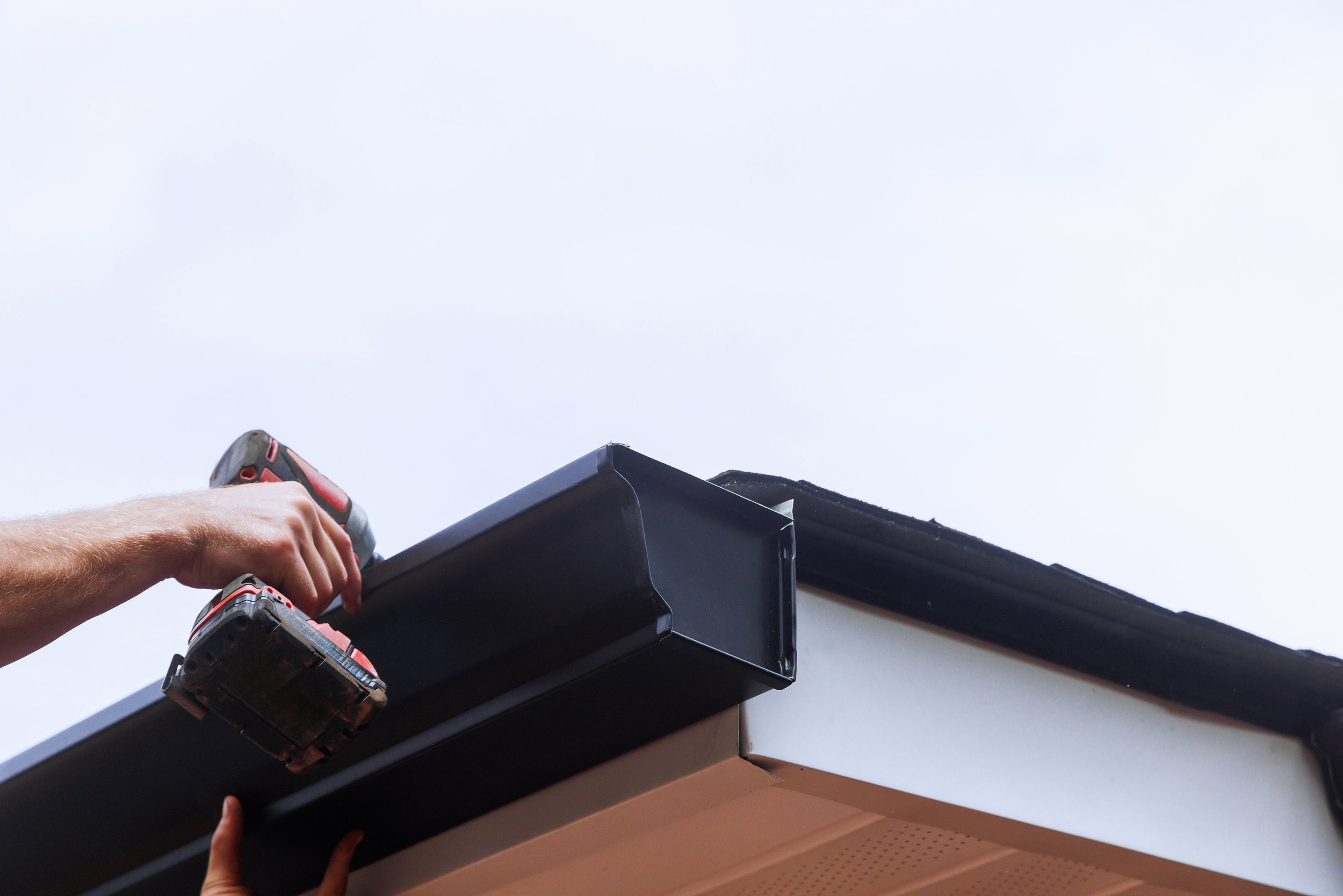 Person using a cordless drill to install or repair the gutter of a house under a cloudy sky.
