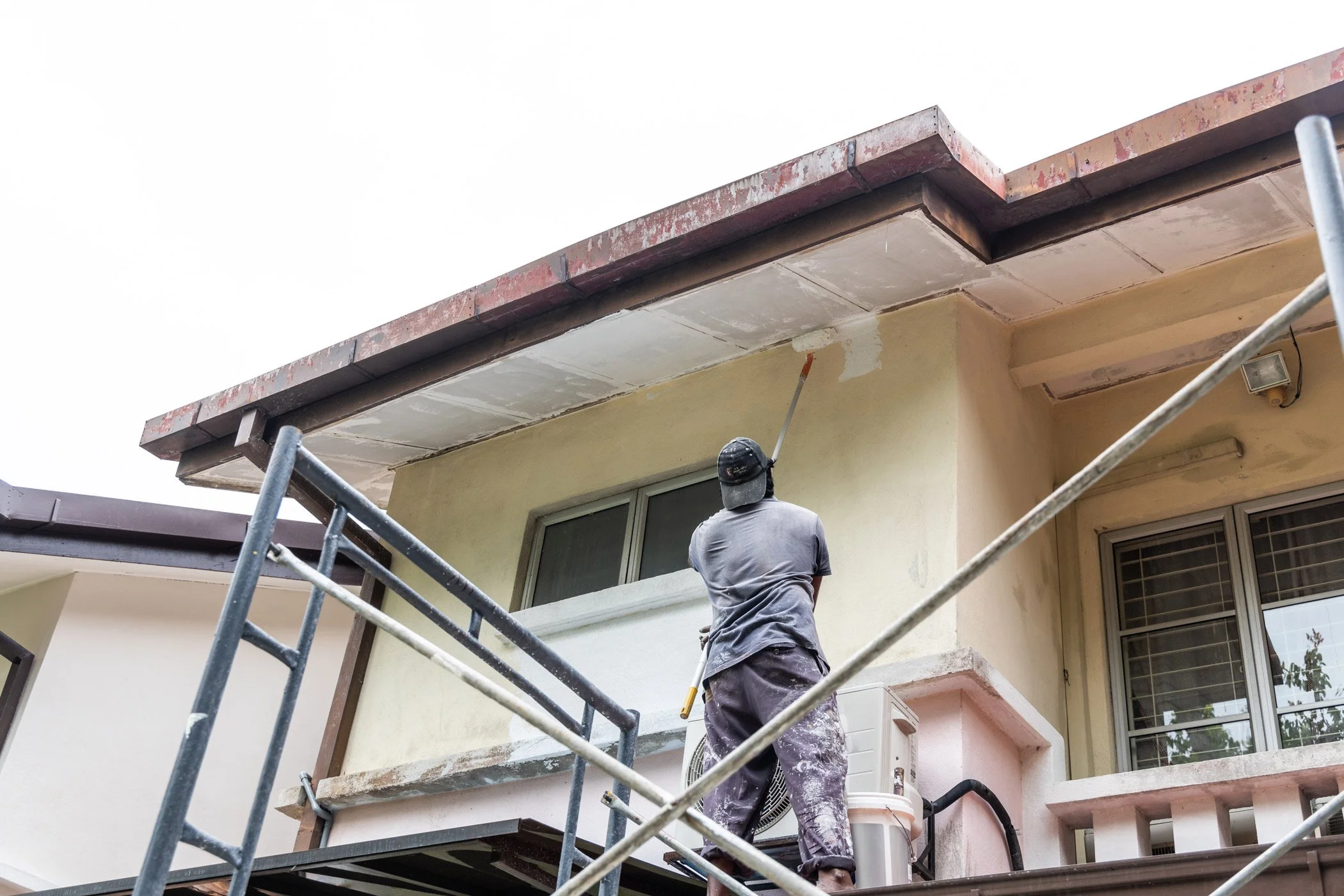 A construction worker in a gray t-shirt and cap is working on the exterior of a house, standing on scaffolding near the roofline.