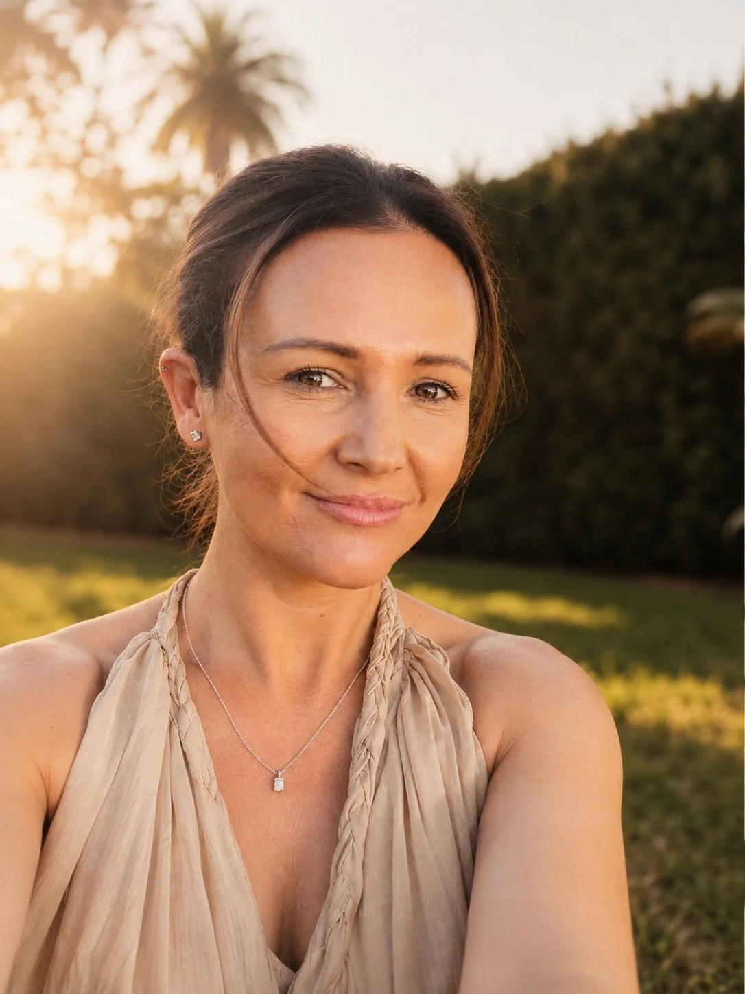 A woman smiling outdoors during sunset, wearing a beige sleeveless top and jewelry, with trees and a palm tree in the background.