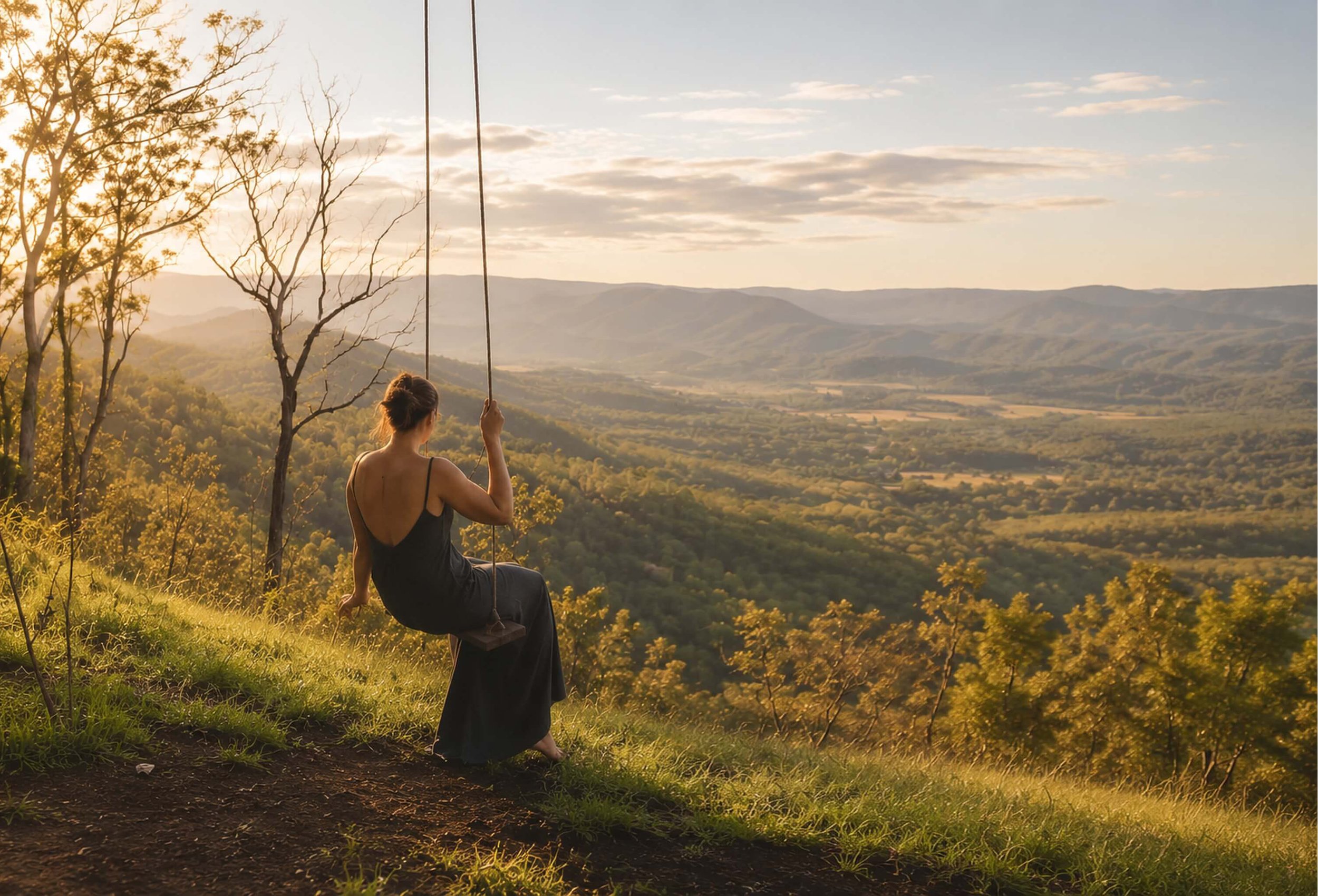 A woman sitting on a swing on a grassy hill at sunset, overlooking a valley with distant hills and a partly cloudy sky.