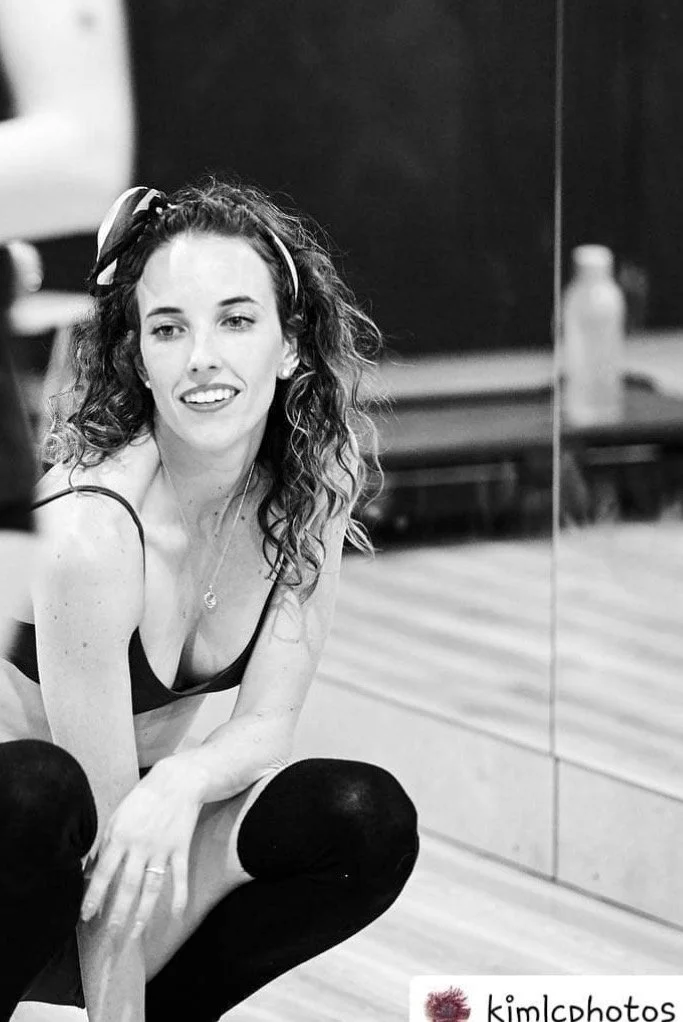 A young woman with curly hair, wearing a headband, sitting in a dance studio next to a mirror, smiling. She is dressed in dance attire, with black tights and a tank top.