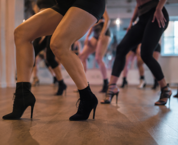 Close-up of women dancing at a studio, wearing high heels and fitted workout clothes, with other dancers in the background.